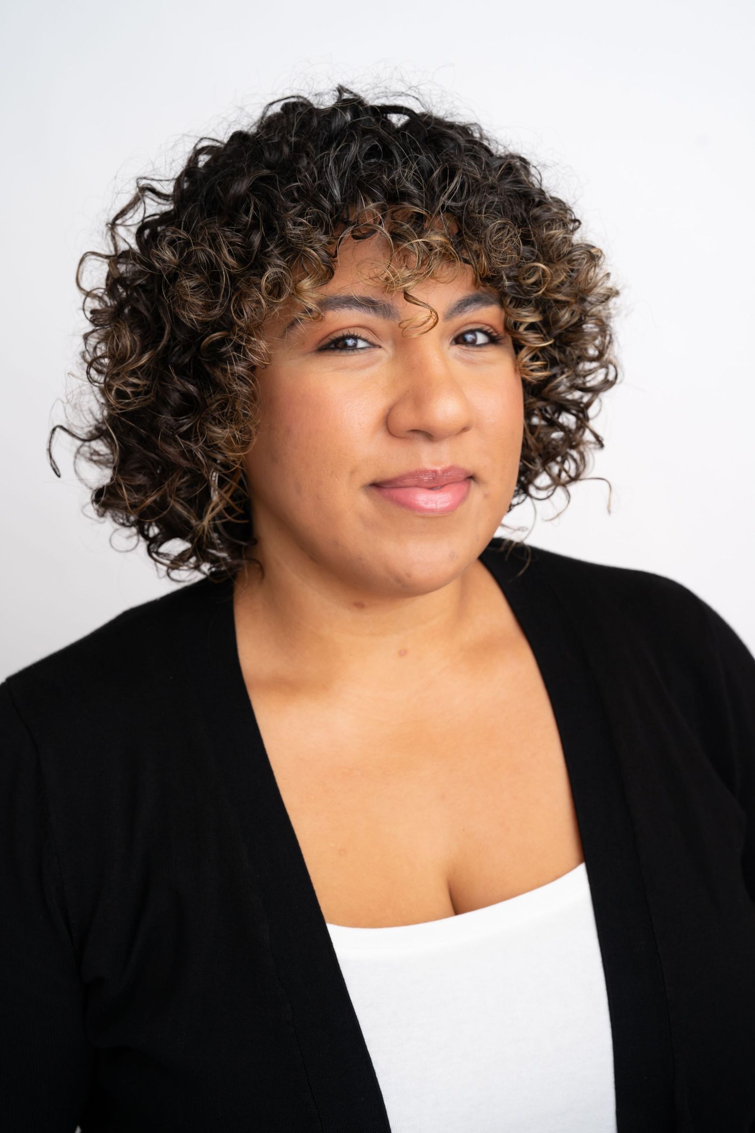 Woman with curly highlighted hair wearing black cardigan and white top, smiling softly.