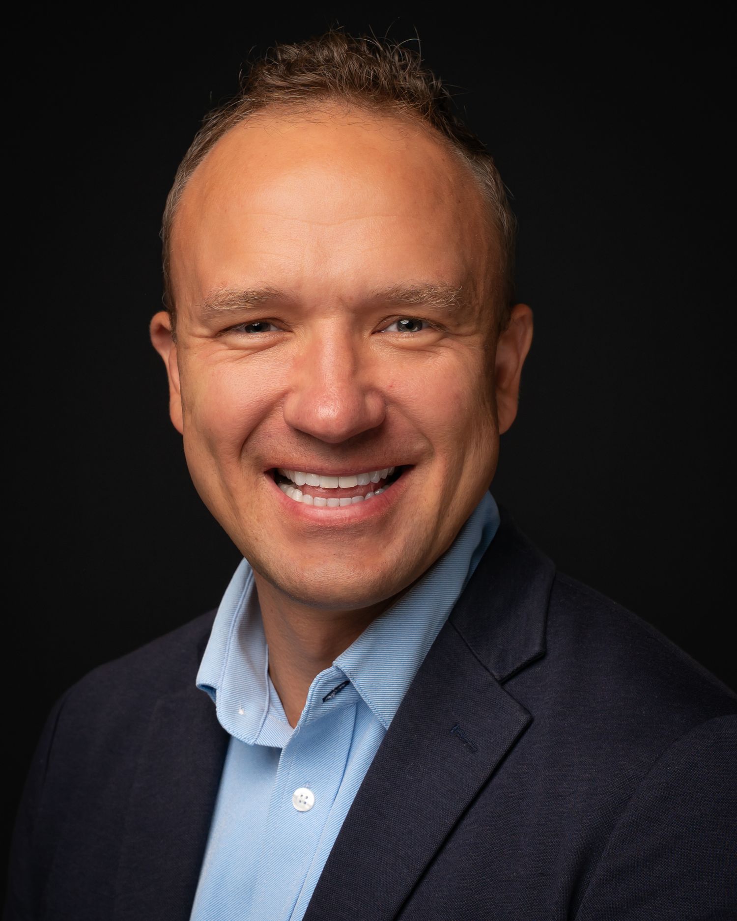 Smiling middle-aged man with short hair wearing a navy blazer and light blue shirt.