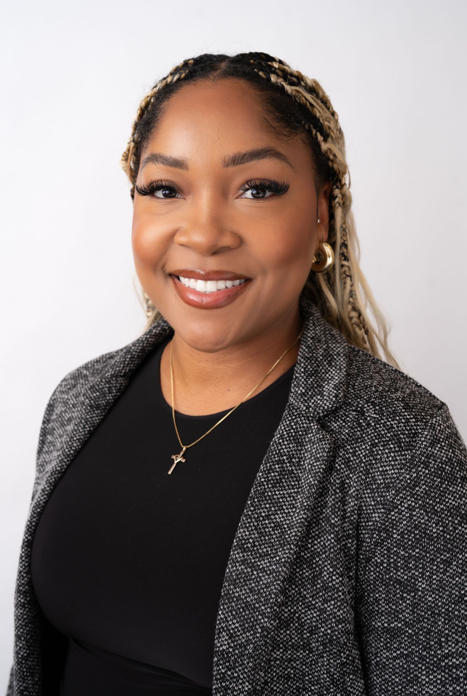 Professional headshot of a smiling woman in a gray blazer and black top, wearing a cross necklace and braided hair.
