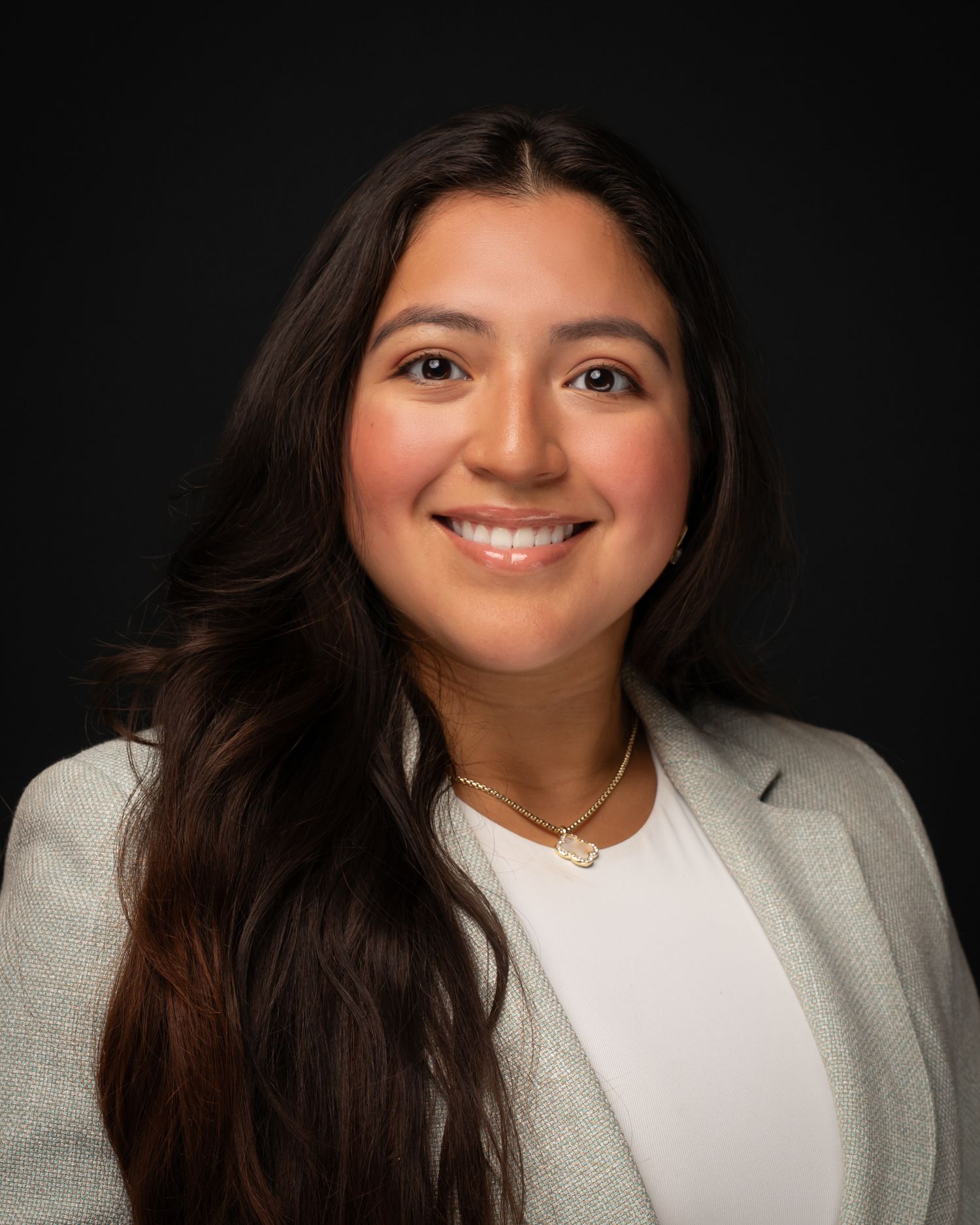 Professional headshot of a young woman with long dark hair, wearing a blazer and gold necklace, smiling confidently.