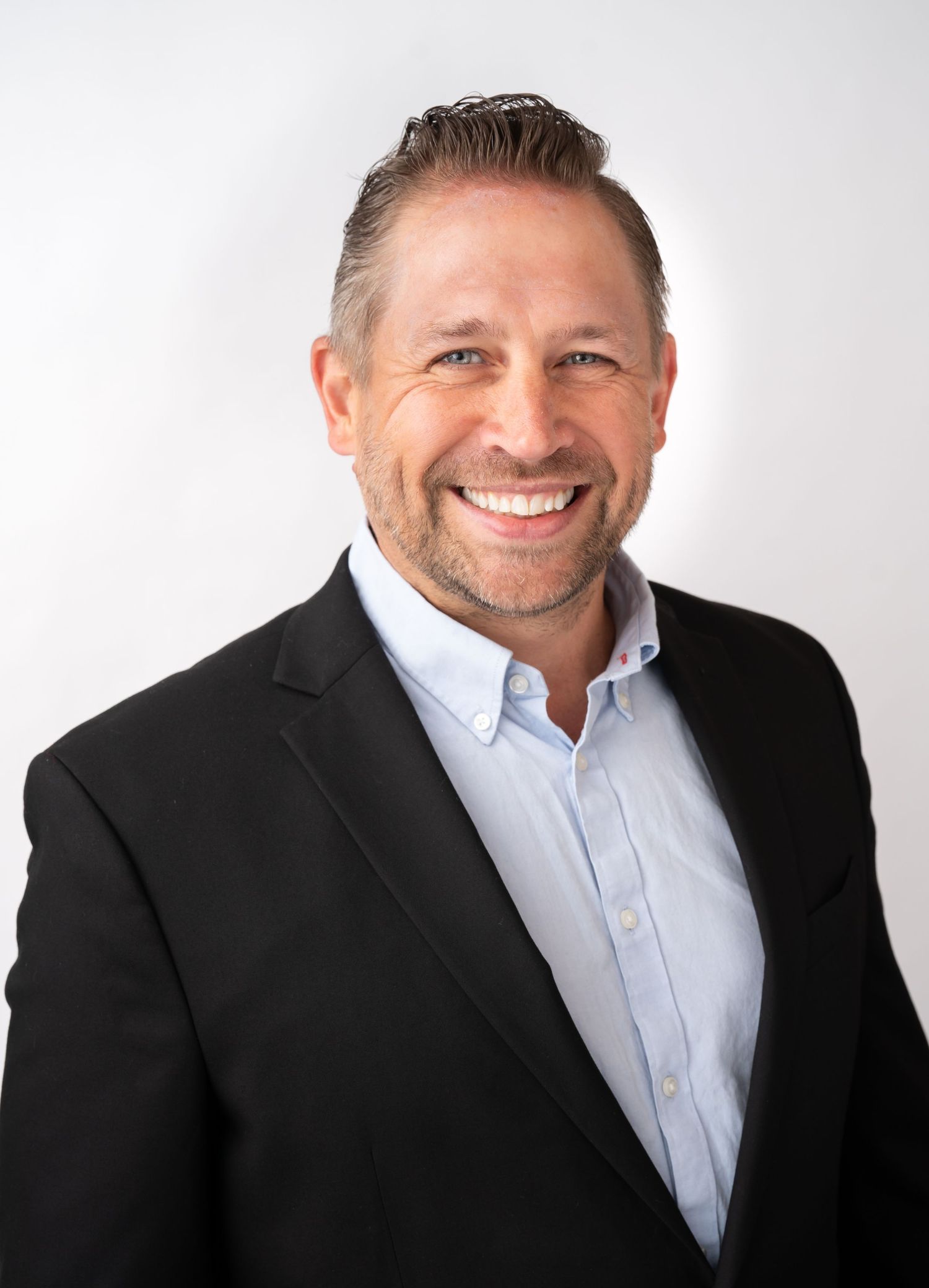 Professional headshot of a smiling middle-aged man in a black blazer and light blue shirt against a white background.