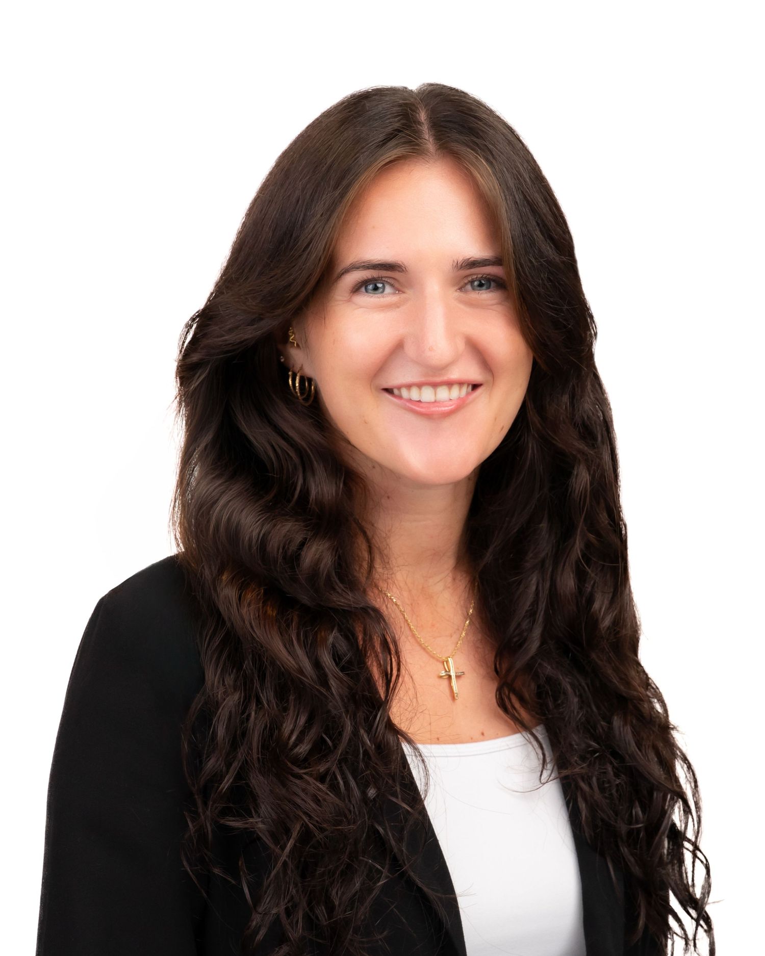 Professional headshot of a smiling young woman with long dark wavy hair wearing a black blazer and gold cross necklace.