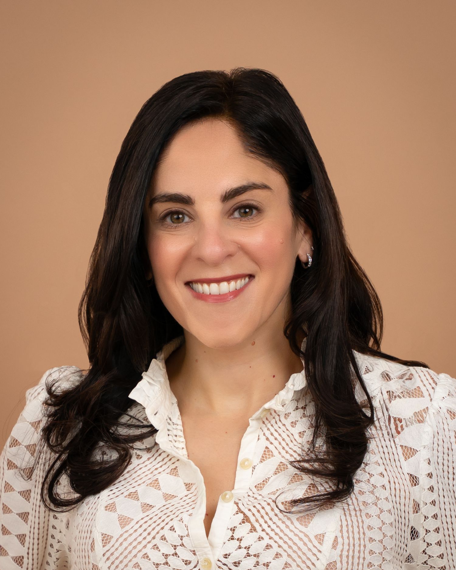 Professional headshot of a smiling woman with dark hair wearing a white lace top against a beige background.