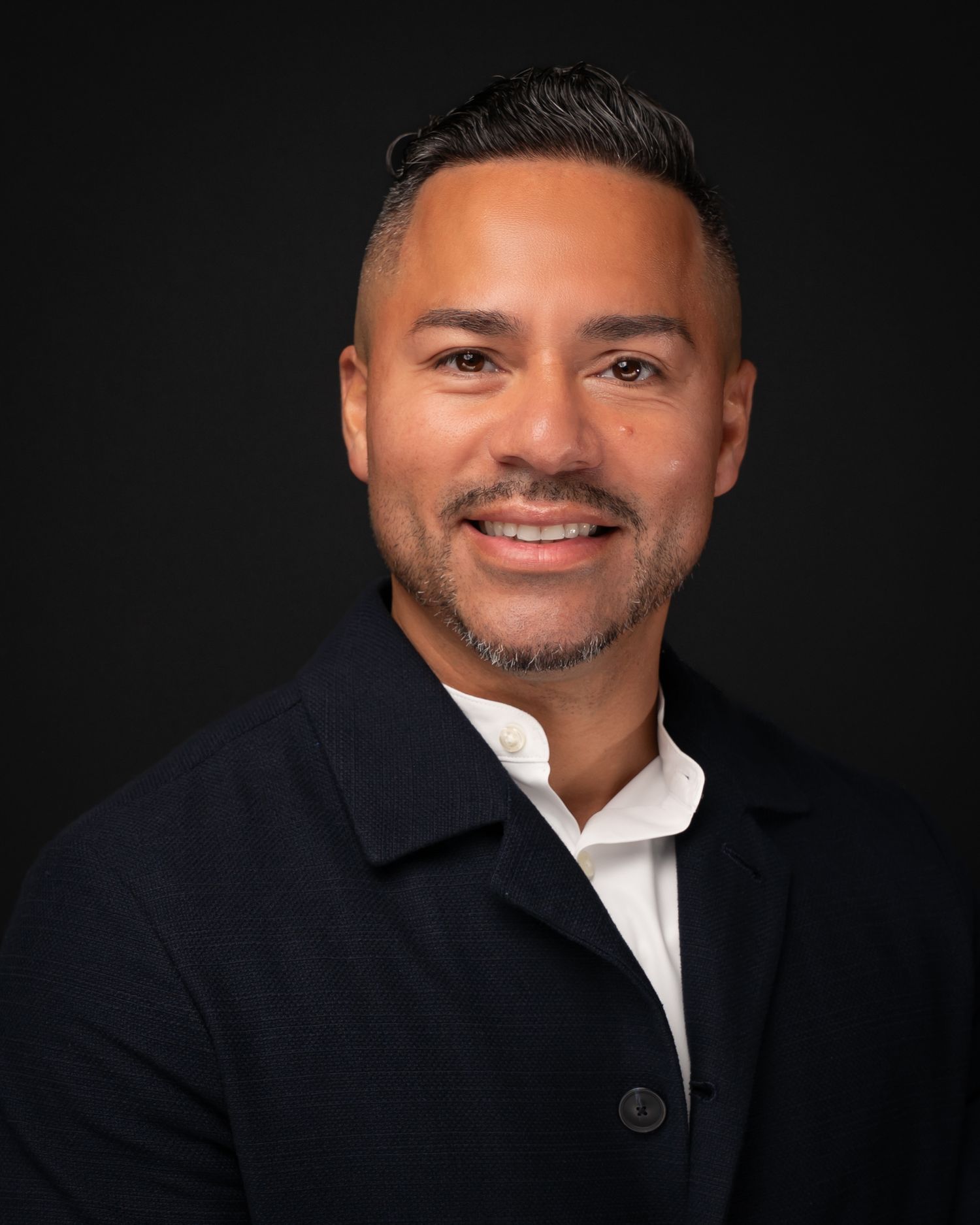 Professional headshot of a smiling man in a dark blazer and white shirt against a black background.