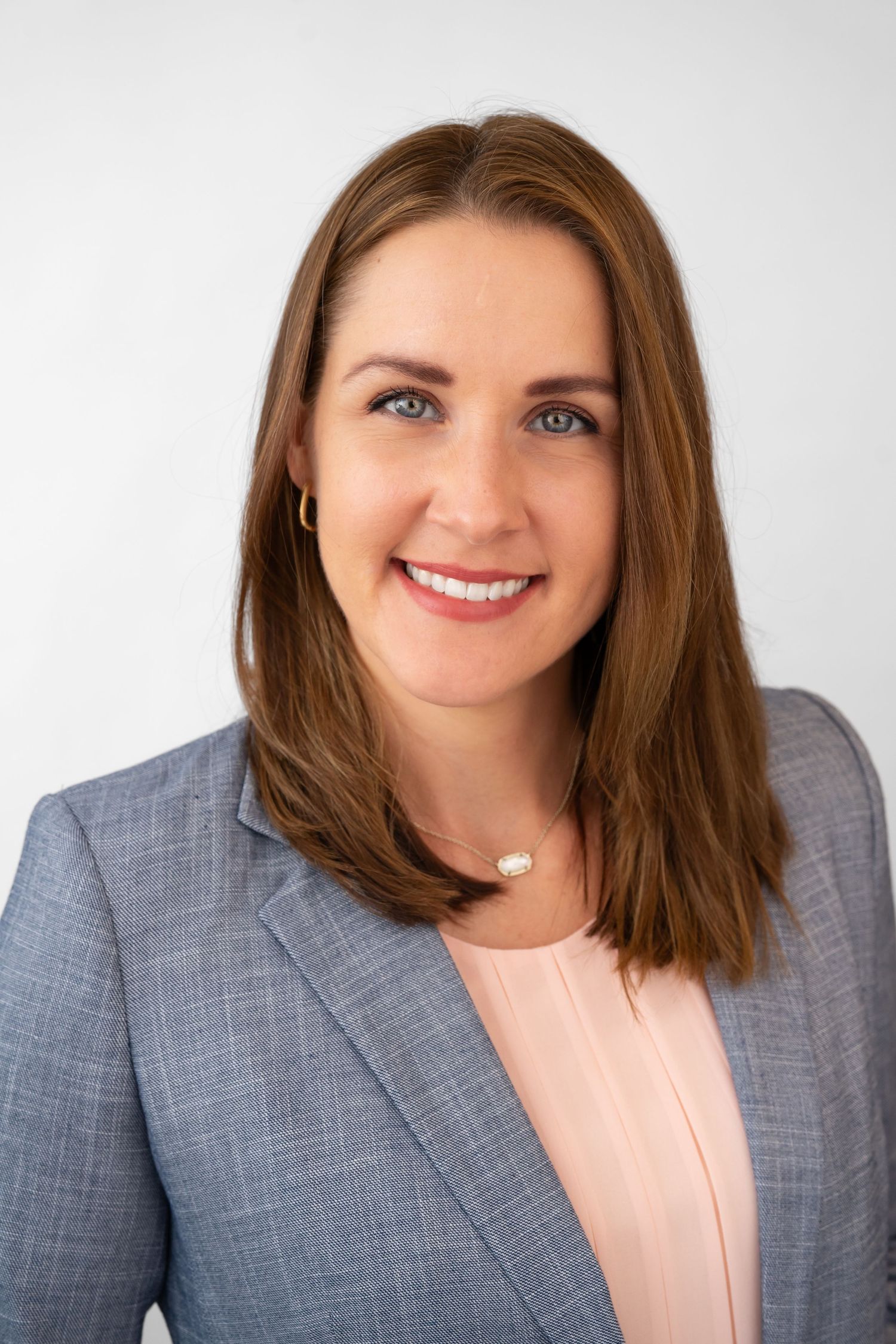Professional headshot of a smiling woman with brown hair in a gray blazer and pearl necklace against a white background.