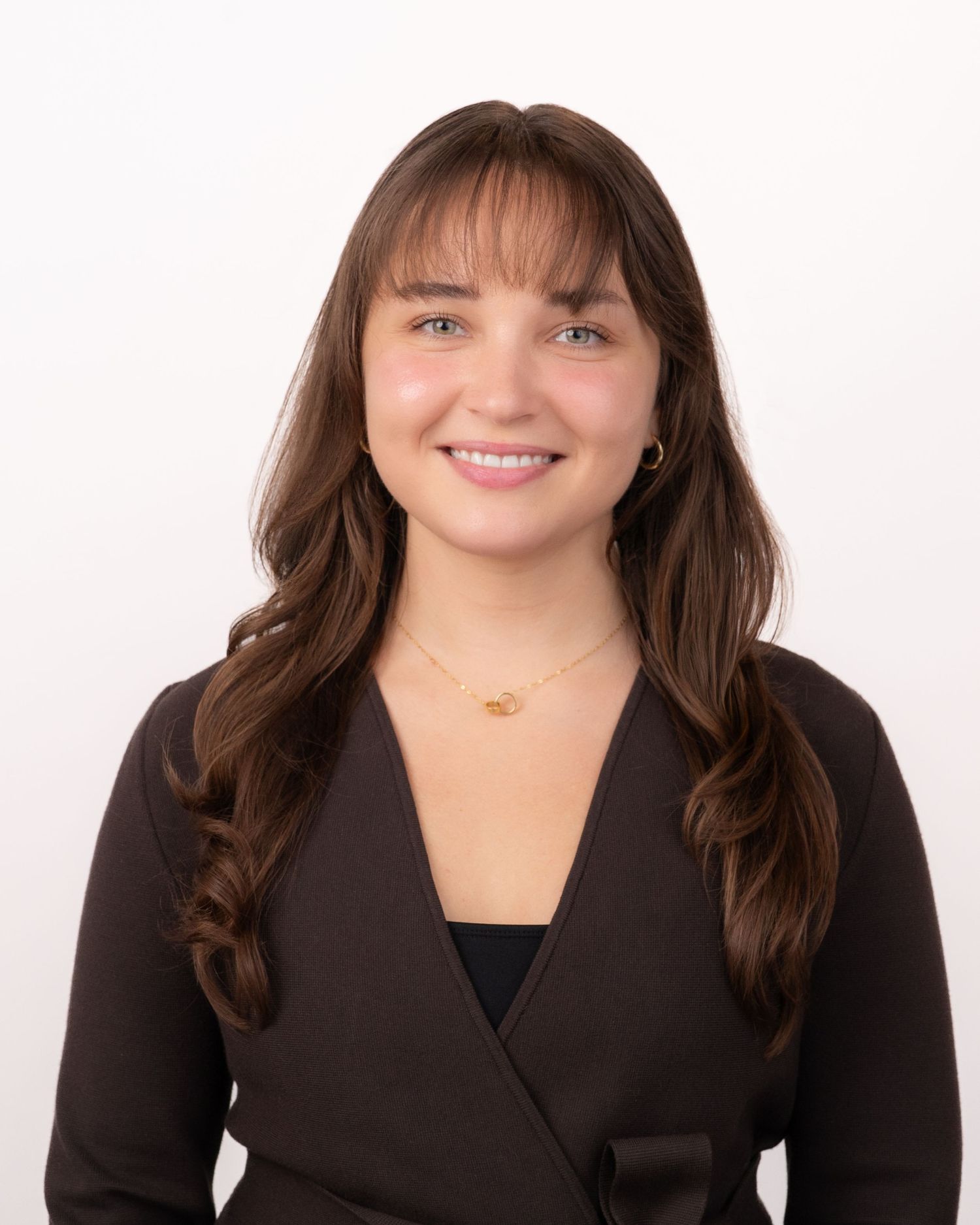 Young woman with long brown hair and bangs smiling in a dark blazer against a white background.