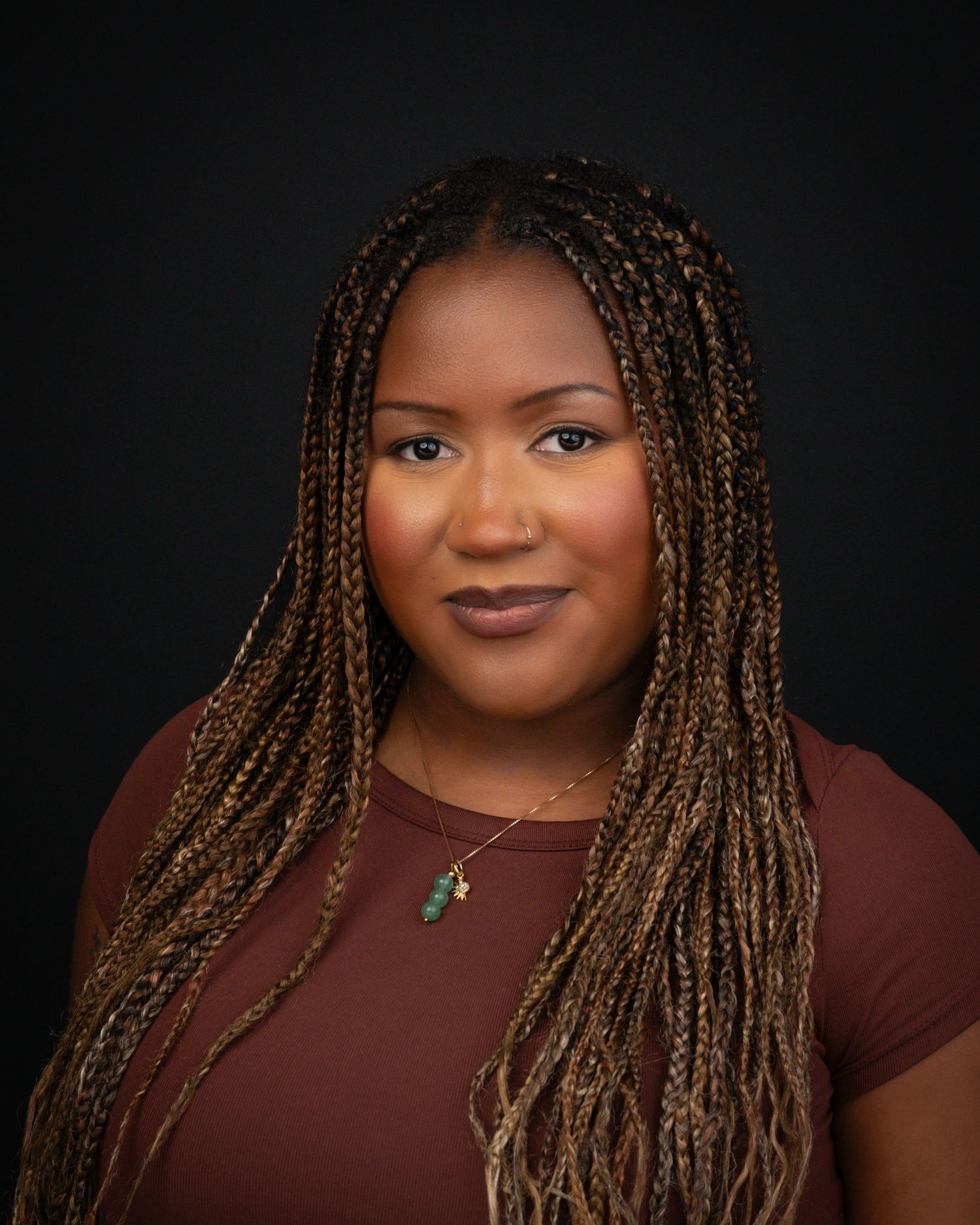 Professional headshot of a woman with long brown box braids wearing a maroon top and delicate necklace against dark background.