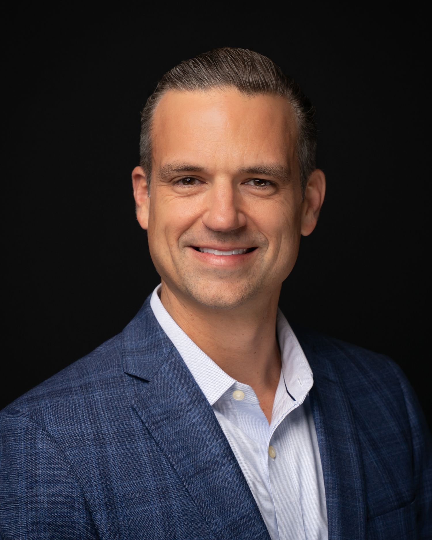 Professional headshot of a smiling middle-aged man in a blue plaid blazer and white shirt against a dark background.