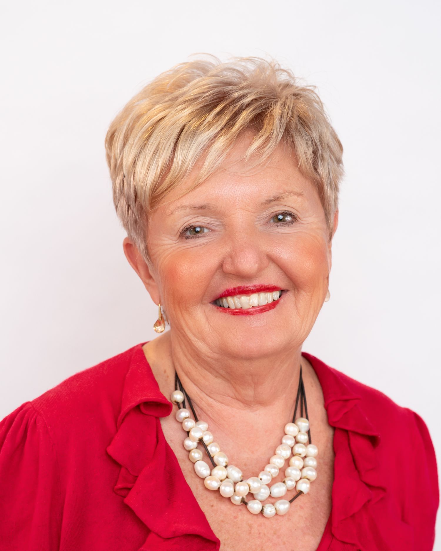 Smiling older woman with short blonde hair wearing a red blouse and pearl necklace against a white background.