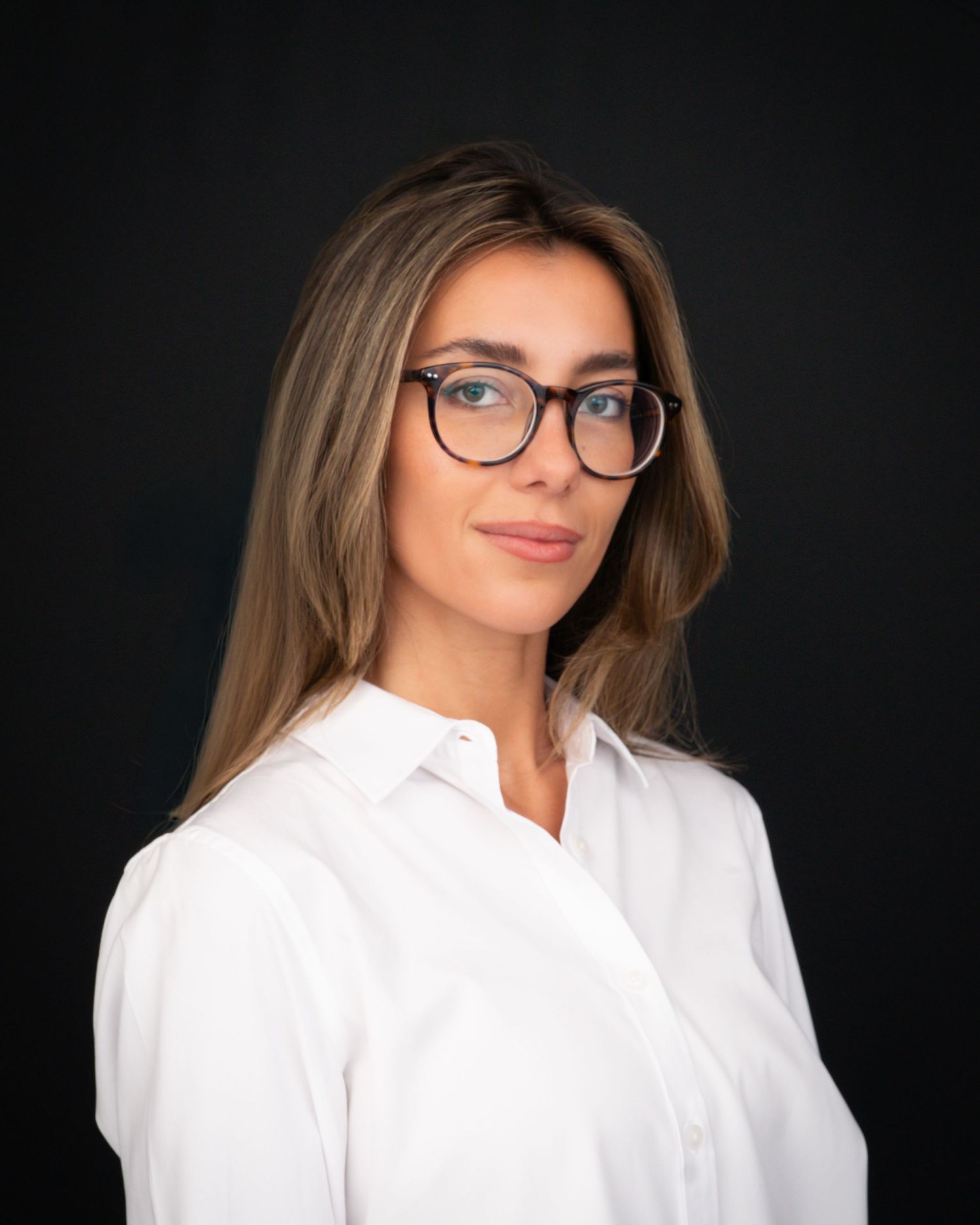 Professional headshot of a young woman with long blonde hair, glasses, and a white button-up shirt against a dark background.