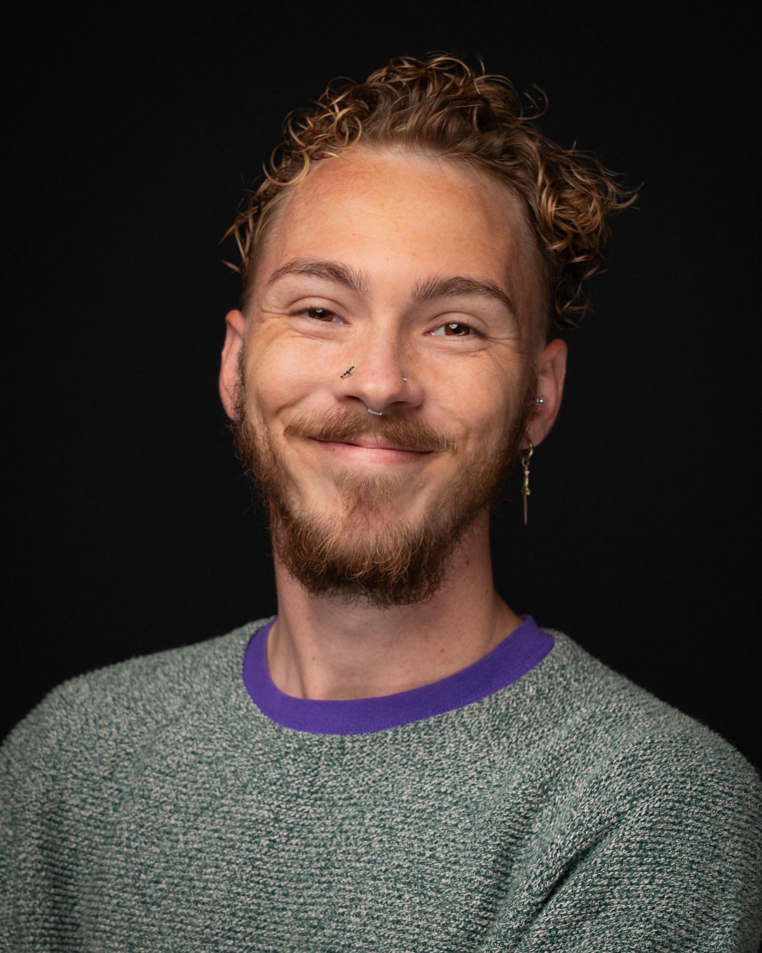Young man with curly blonde hair, beard, nose ring, and earring smiling in a green sweater with purple collar.