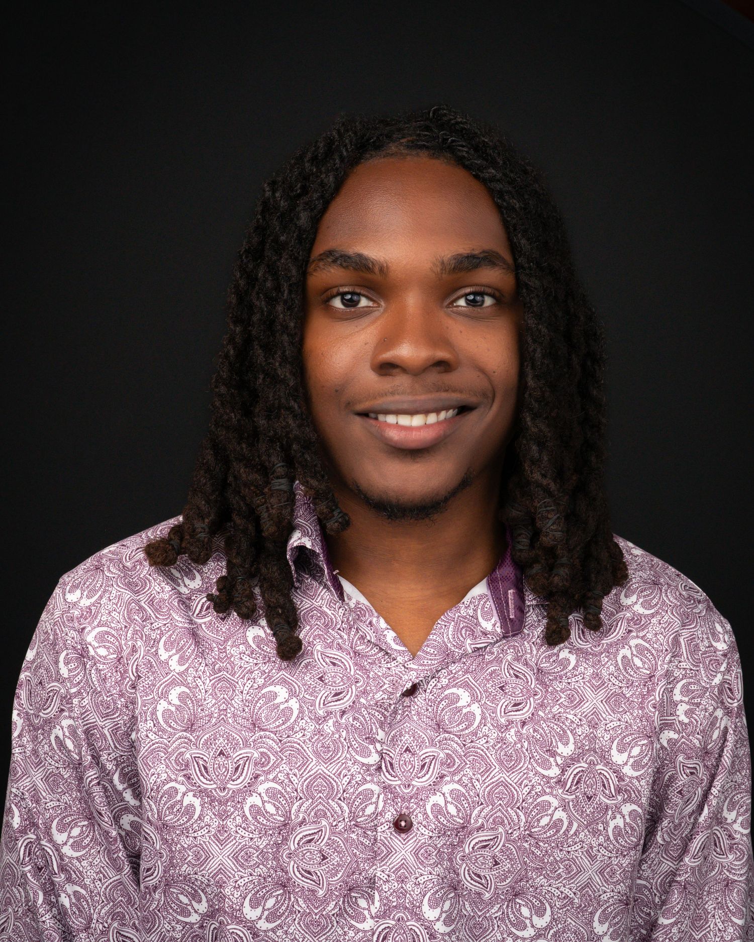 Young man with locs wearing a patterned purple shirt, smiling confidently against a dark background.
