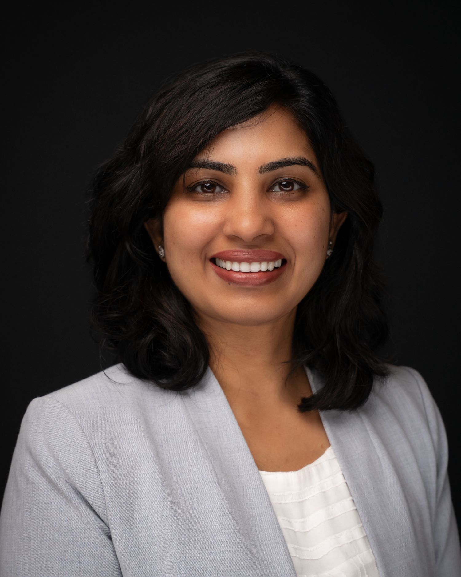 Professional woman in a gray blazer with dark wavy hair, smiling warmly against a dark background.