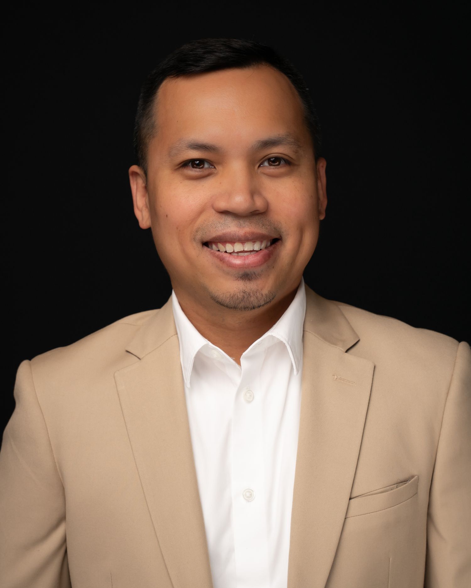 Professional headshot of a smiling man in a beige blazer and white shirt against a dark background.
