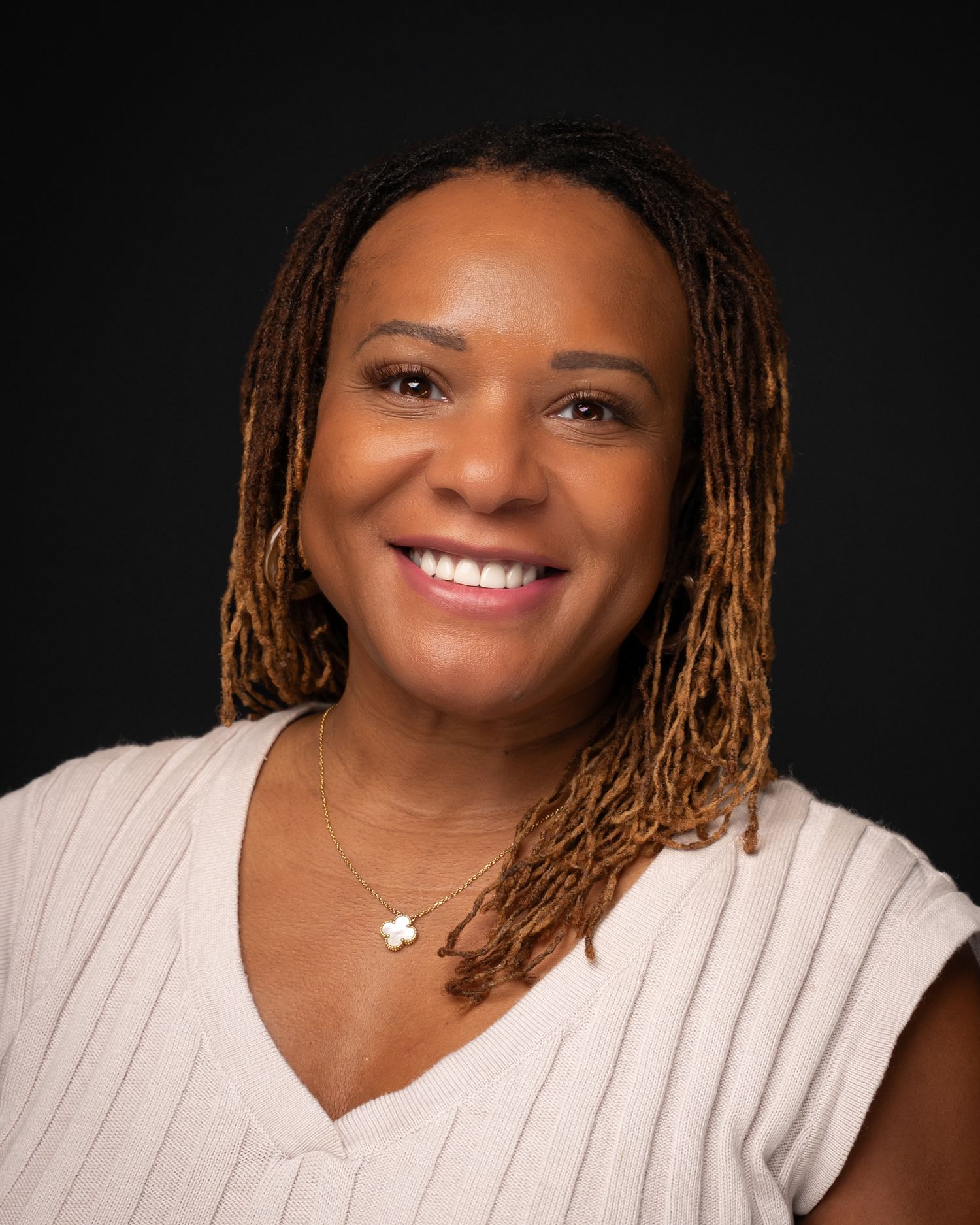 Woman with locs wearing a white sleeveless top and gold necklace, smiling warmly against a dark background.