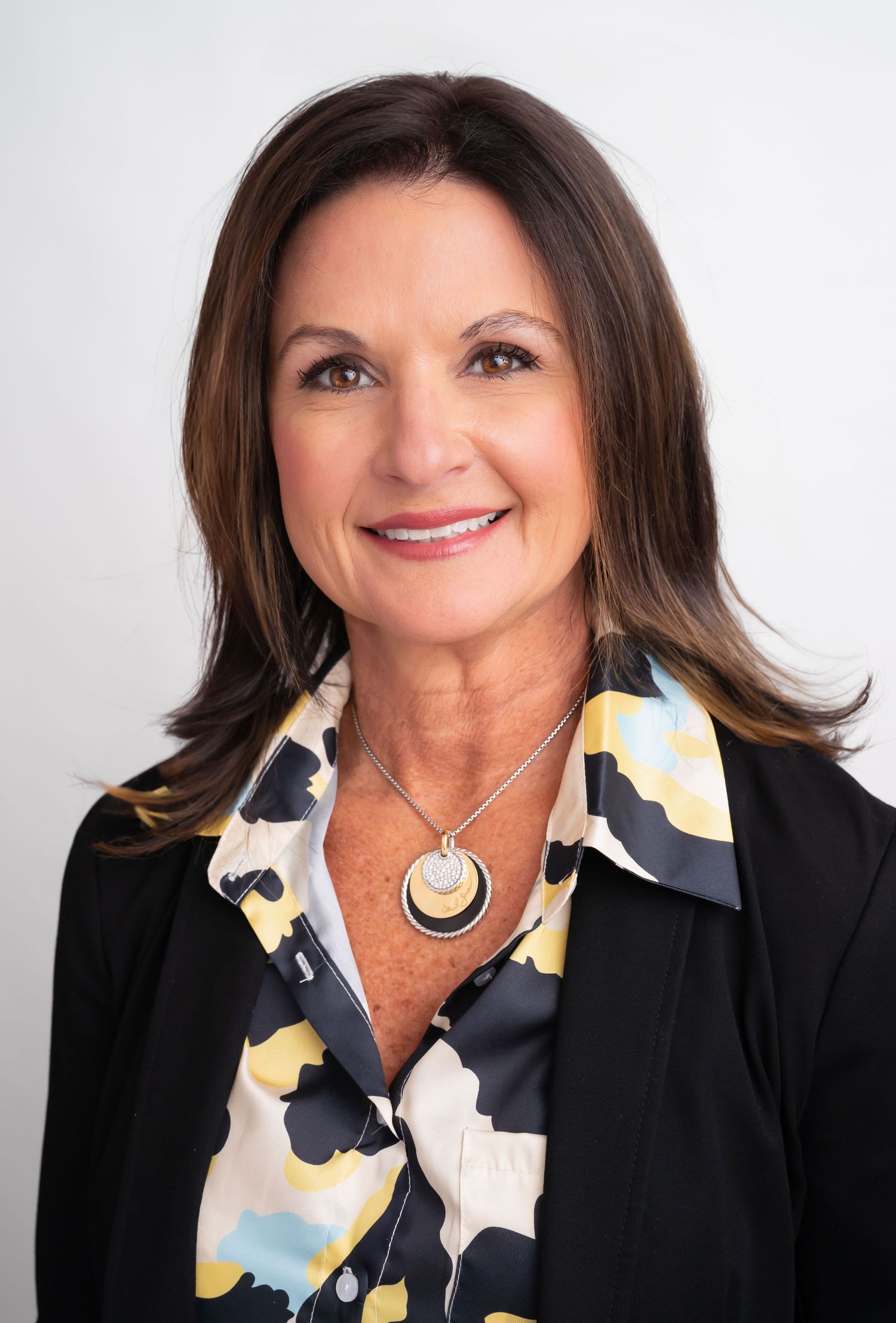 Professional headshot of a smiling woman with dark hair wearing a black blazer, floral blouse, and layered necklace.