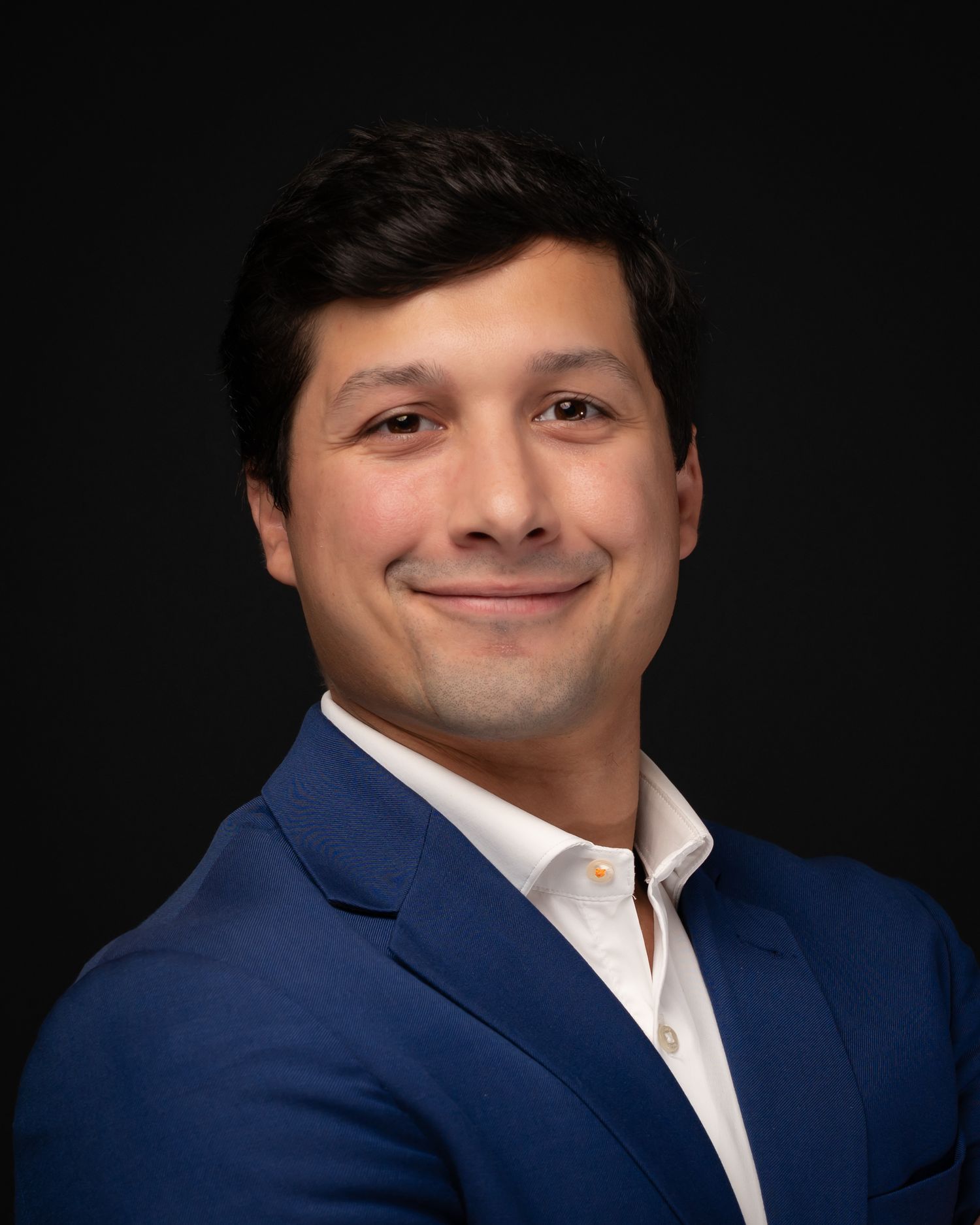 Professional headshot of a smiling young man in a blue blazer and white shirt against a dark background.