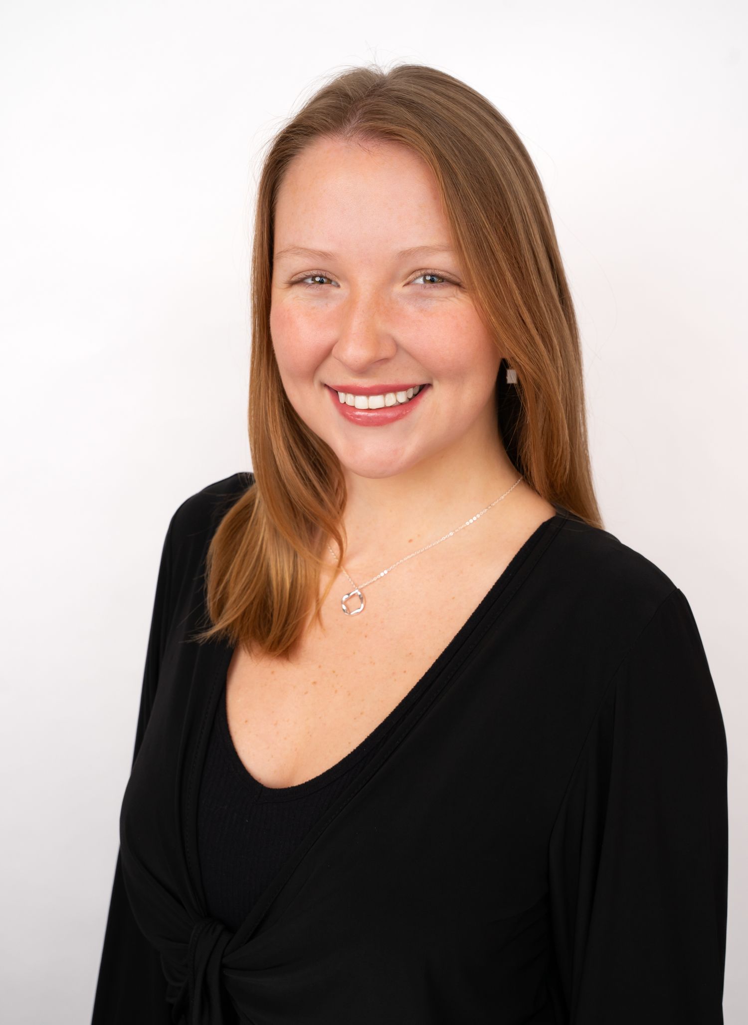 Smiling woman with long auburn hair wearing a black top and delicate necklace against a white background.
