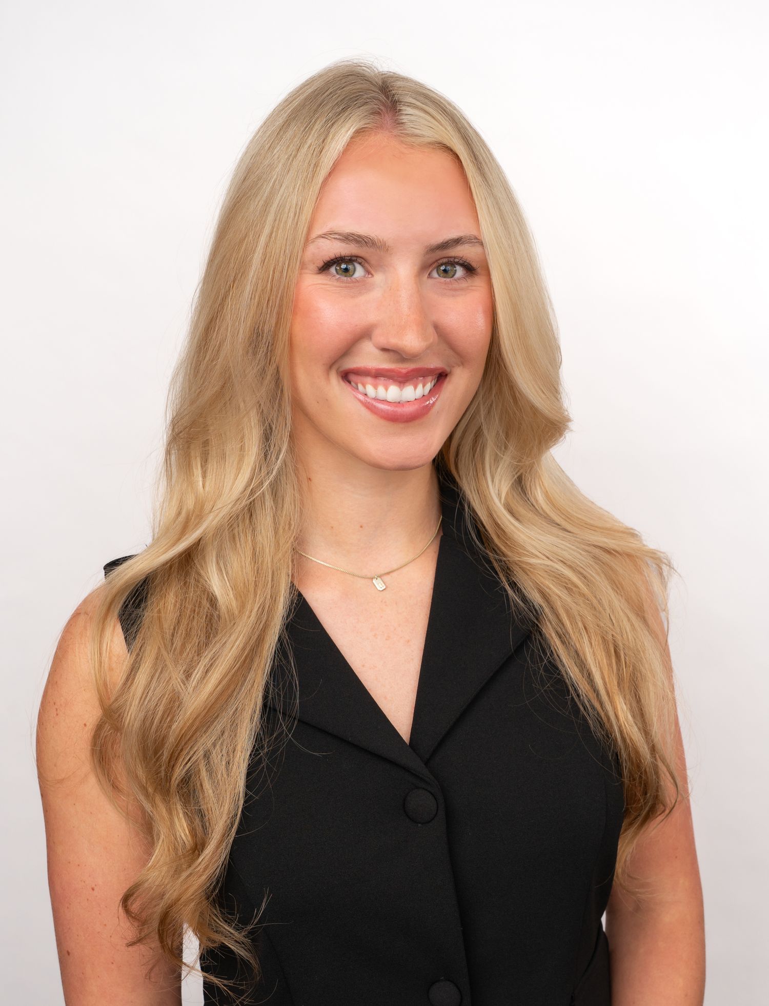 Smiling young woman with long blonde wavy hair wearing a black sleeveless blazer and delicate necklace against white background.
