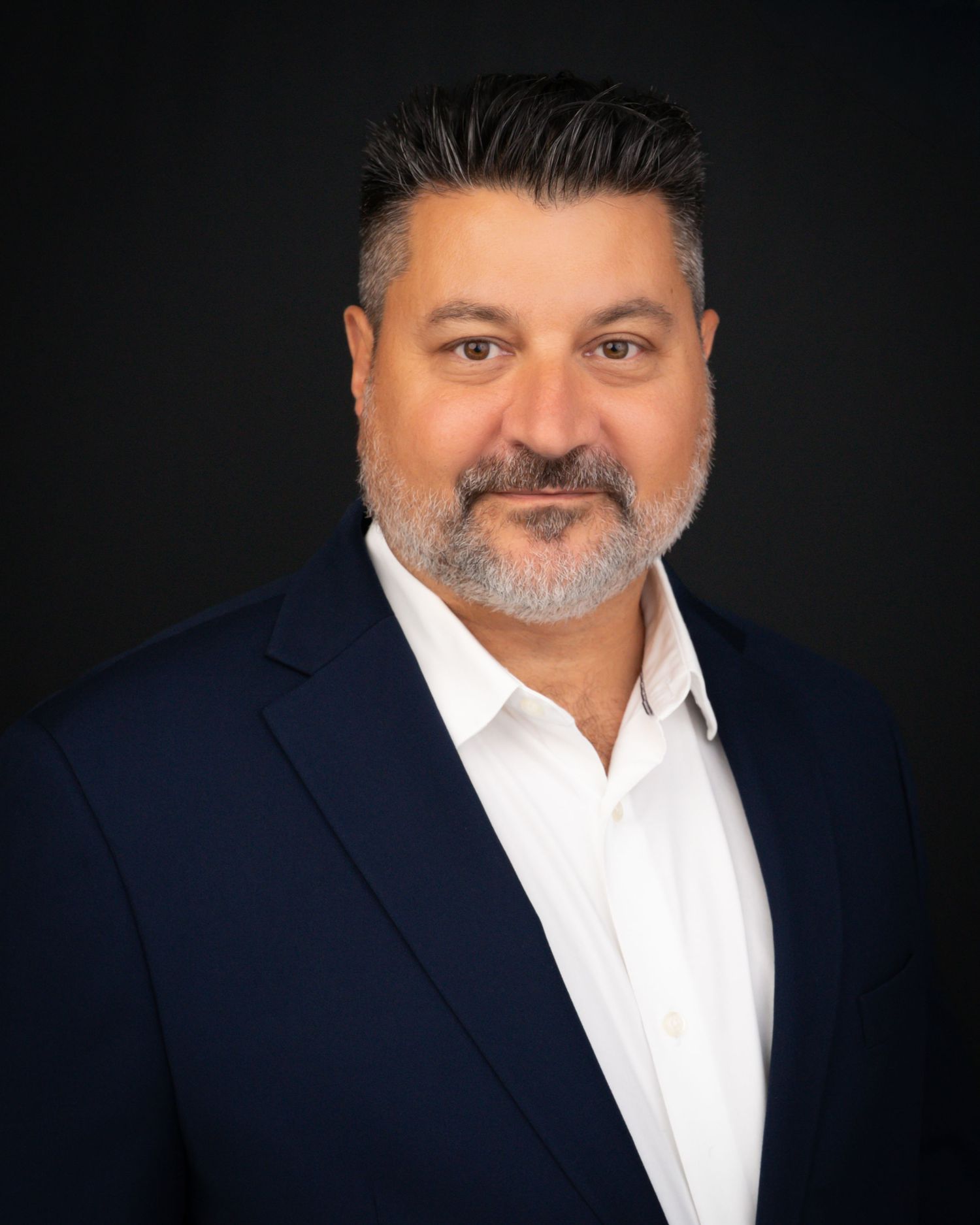 Professional headshot of a man with dark hair and beard wearing a navy blazer and white shirt.