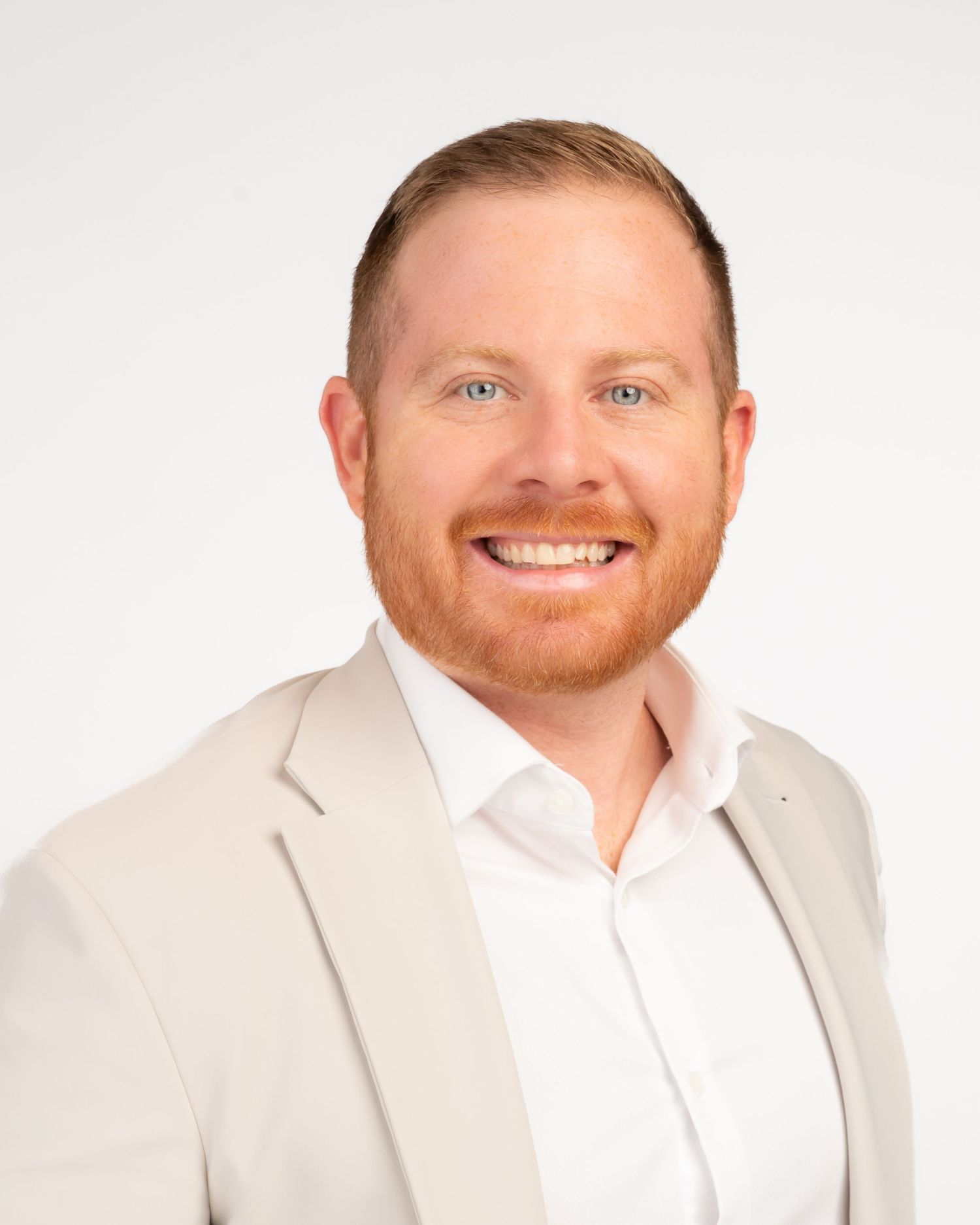 Professional headshot of a smiling red-haired man in a beige blazer and white shirt against a white background.