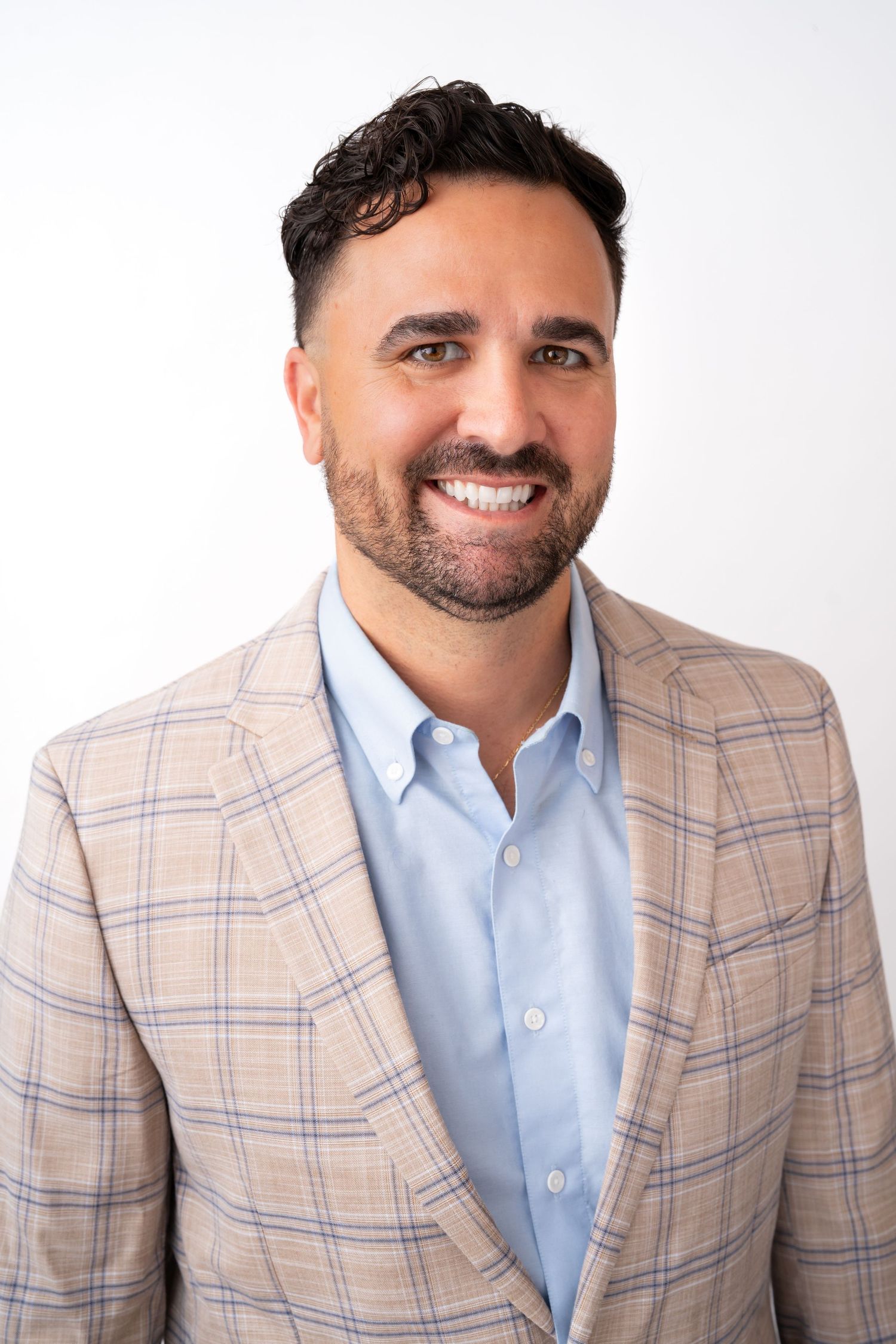Smiling man with dark hair and beard wearing a plaid blazer and light blue shirt against a white background.