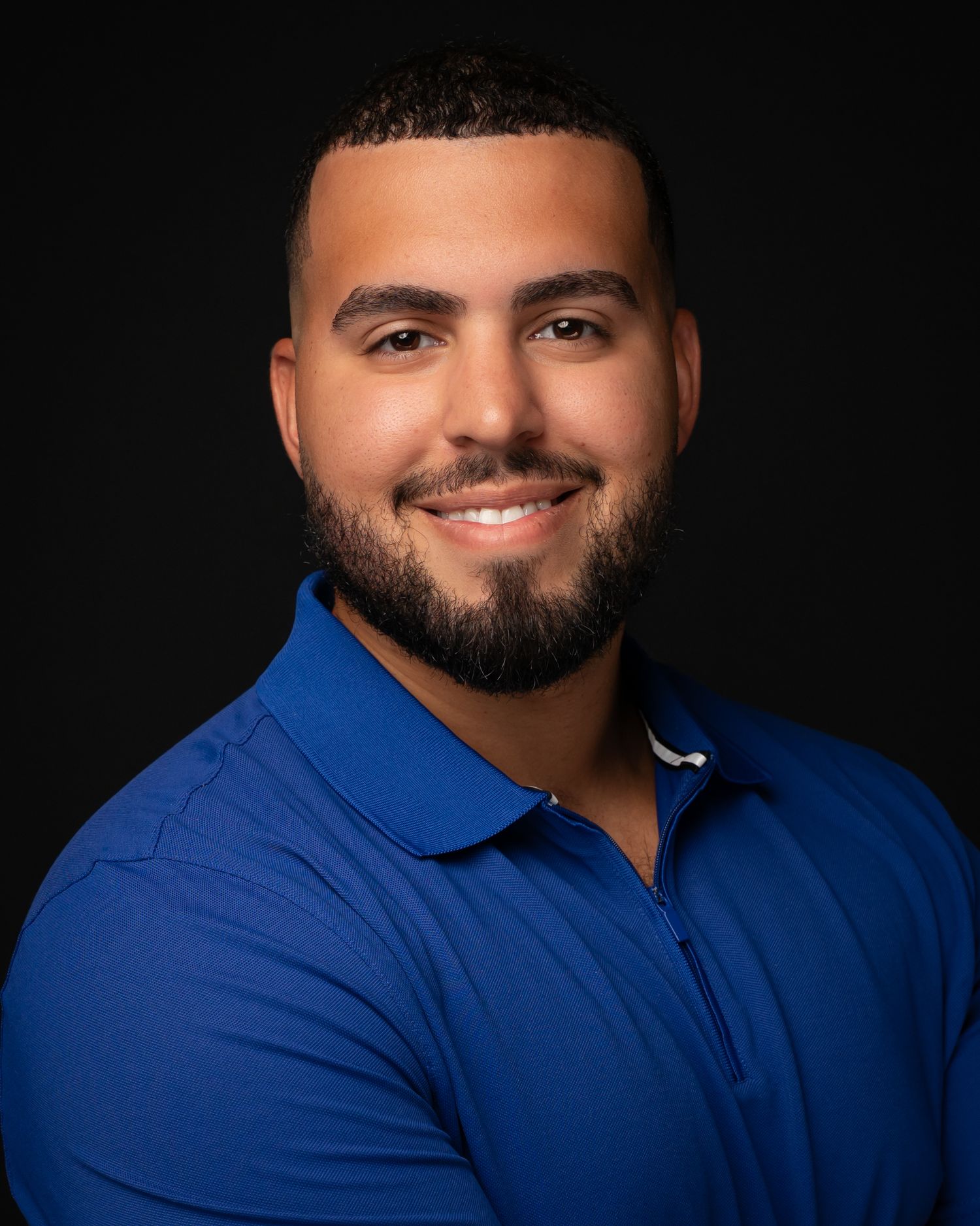 Professional headshot of a smiling man with a beard wearing a blue polo shirt against a dark background.