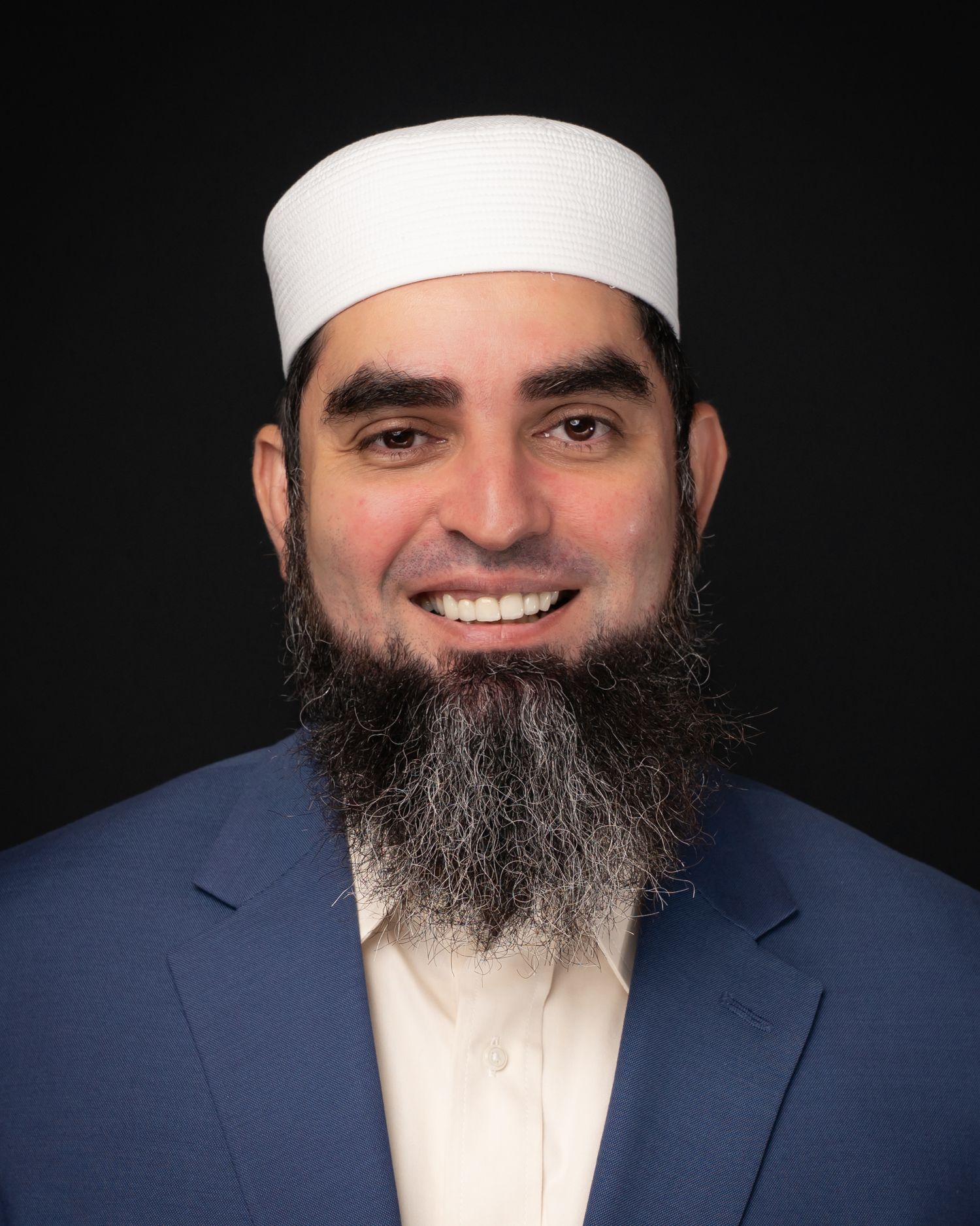Smiling man with a beard wearing a white kufi cap and blue blazer against a dark background.