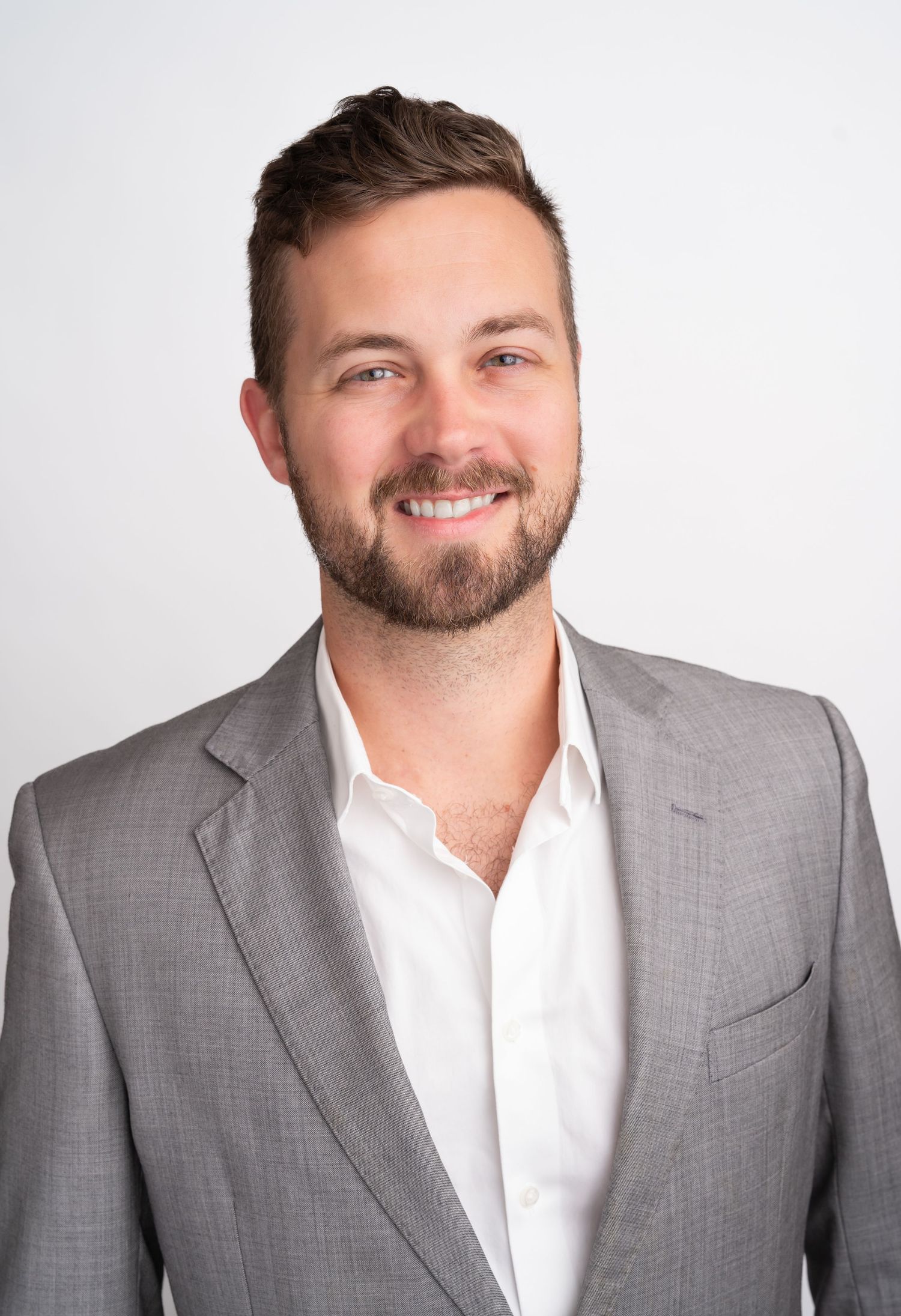 Smiling man with beard wearing gray blazer and white shirt against light background.