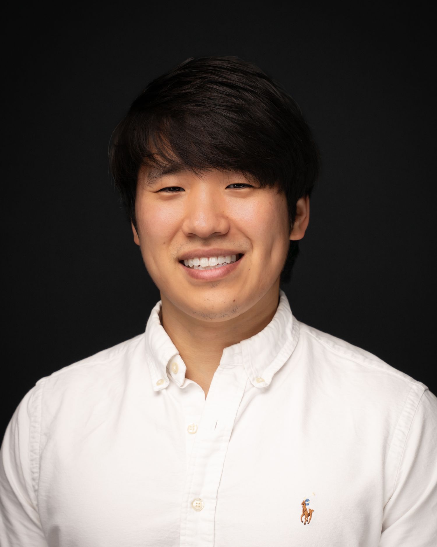 Young man smiling in a white Ralph Lauren button-up shirt against a dark background.