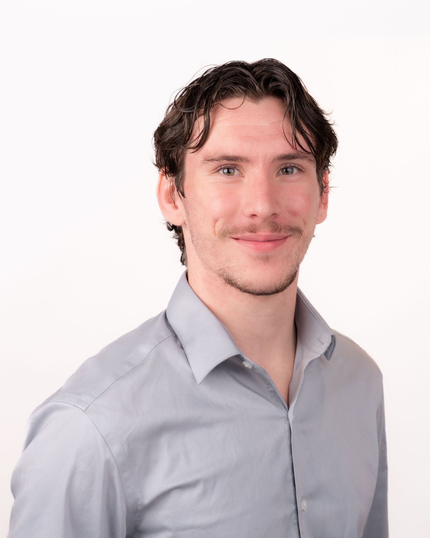 Professional headshot of a young man with dark wavy hair wearing a gray button-up shirt against a white background.
