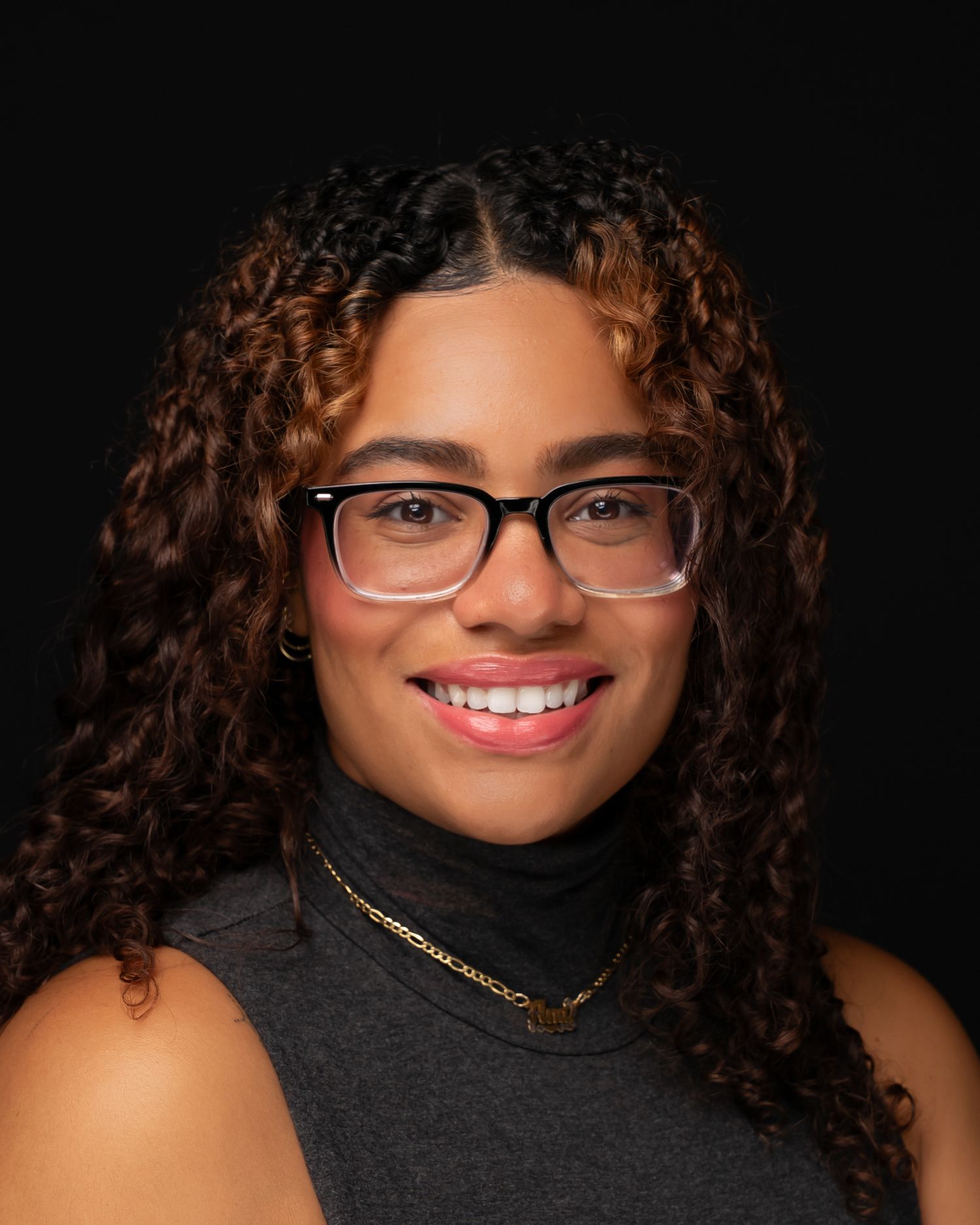 A young woman with curly brown hair and glasses smiles confidently, wearing a gray turtleneck and gold necklace.