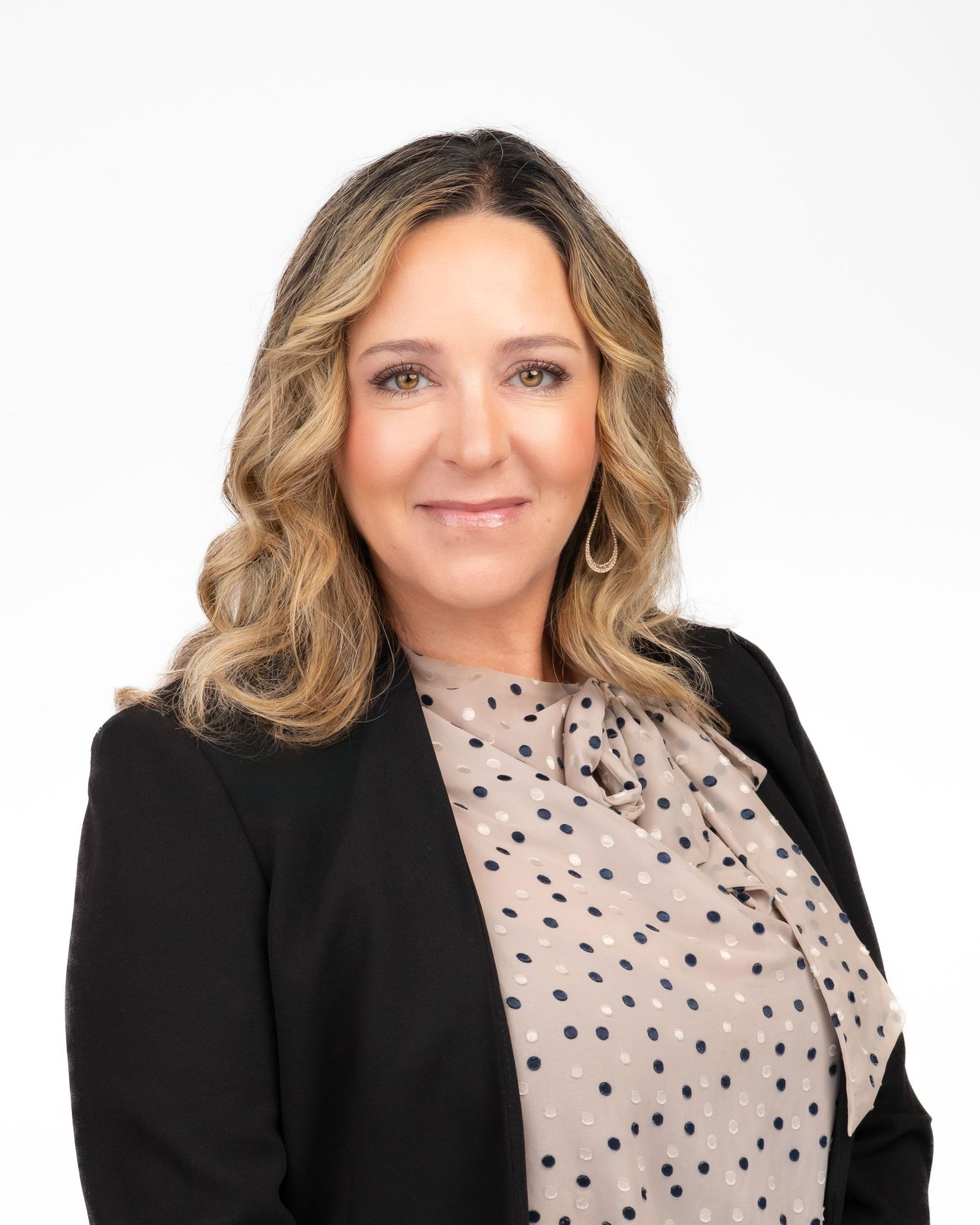 Professional woman with blonde wavy hair wearing a black blazer and polka dot blouse against white background.