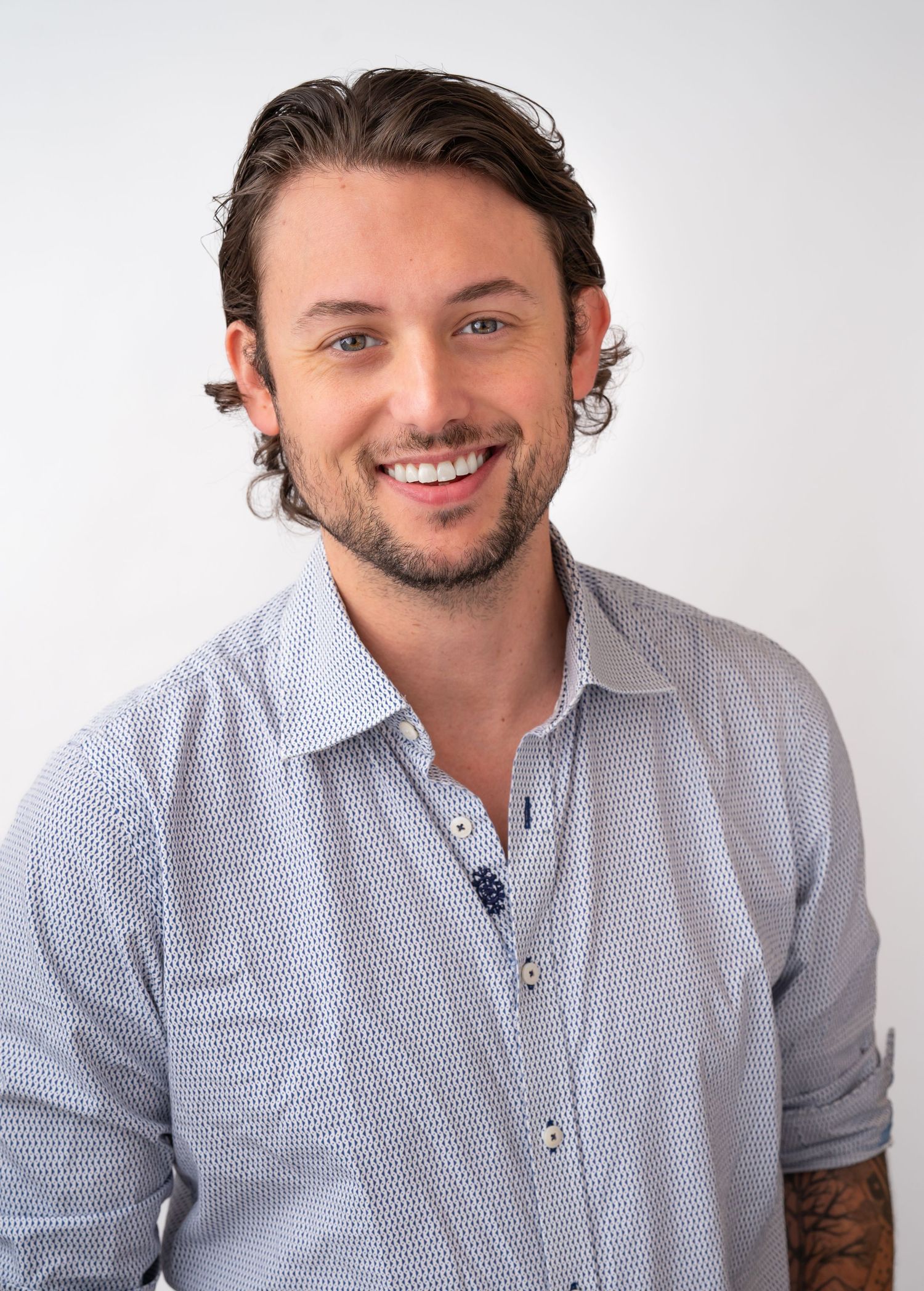 Smiling young man with dark hair and beard wearing a light blue button-up shirt, professional headshot photo.