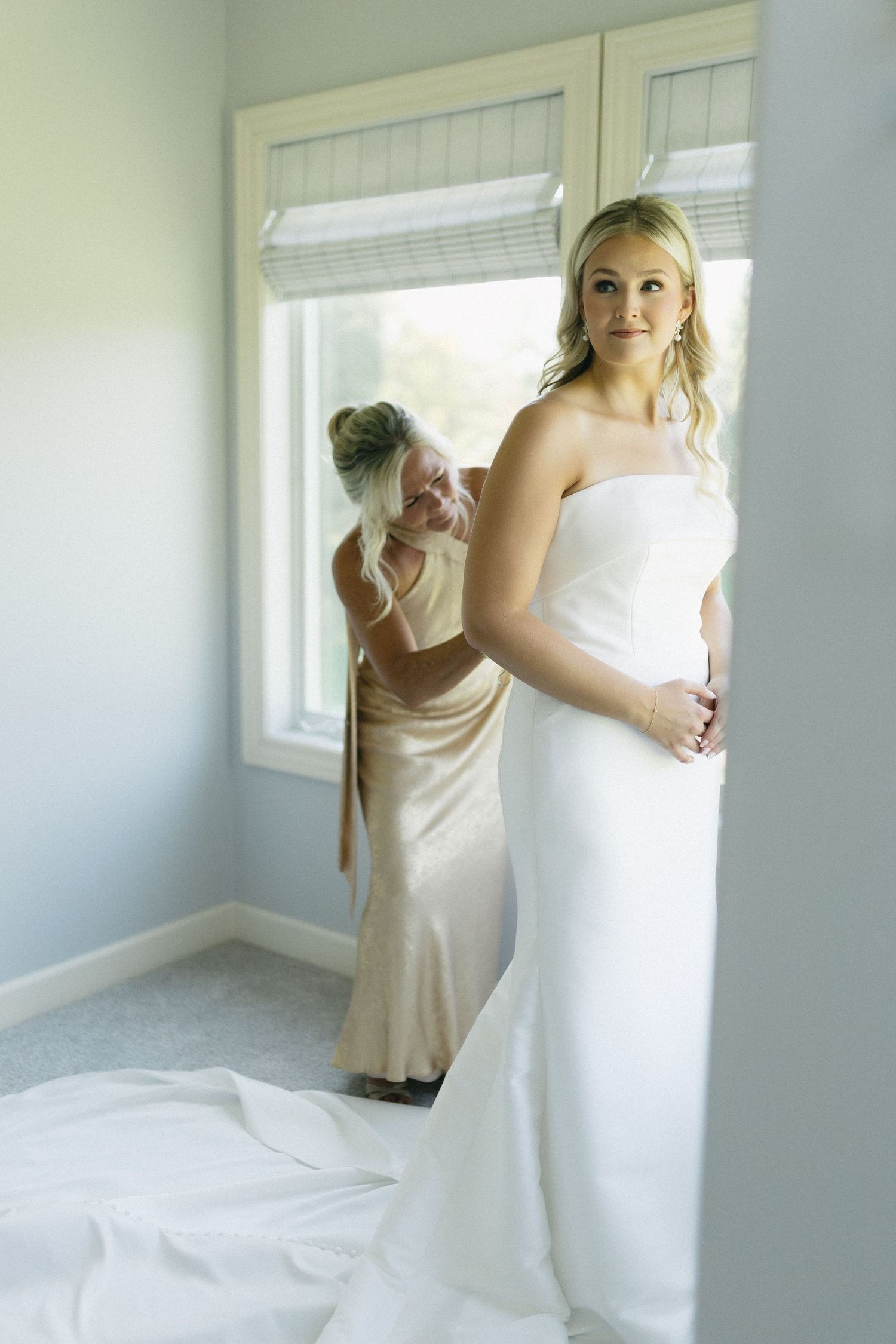Two people in white dresses preparing for a wedding in a bright sunlit room.