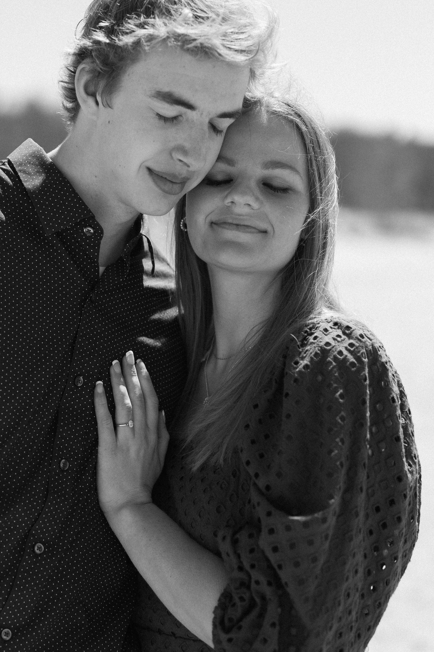 Black and white photo of a young couple embracing tenderly, woman wearing engagement ring, eyes closed in romantic moment.