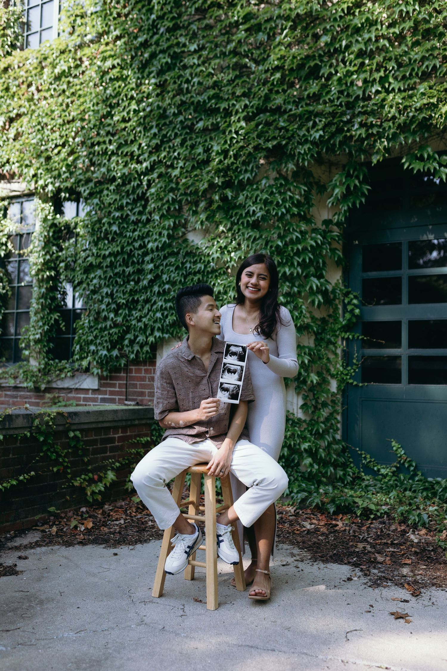 Casual engagement photo of couple sharing a laugh near an ivy-covered brick wall.