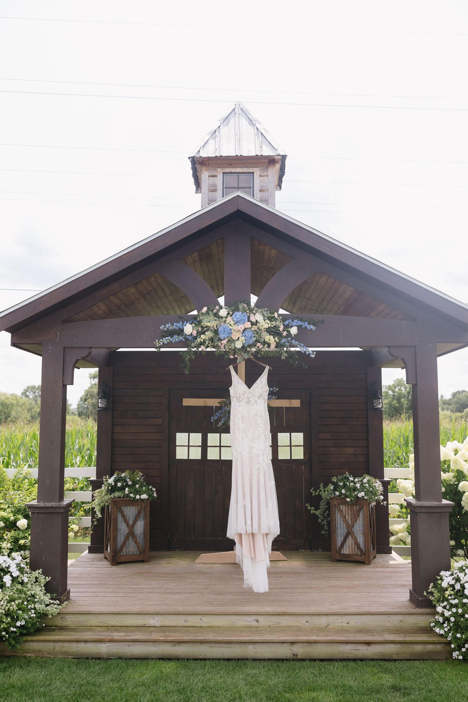 White wedding dress hanging on rustic wooden gazebo decorated with blue and white flowers.