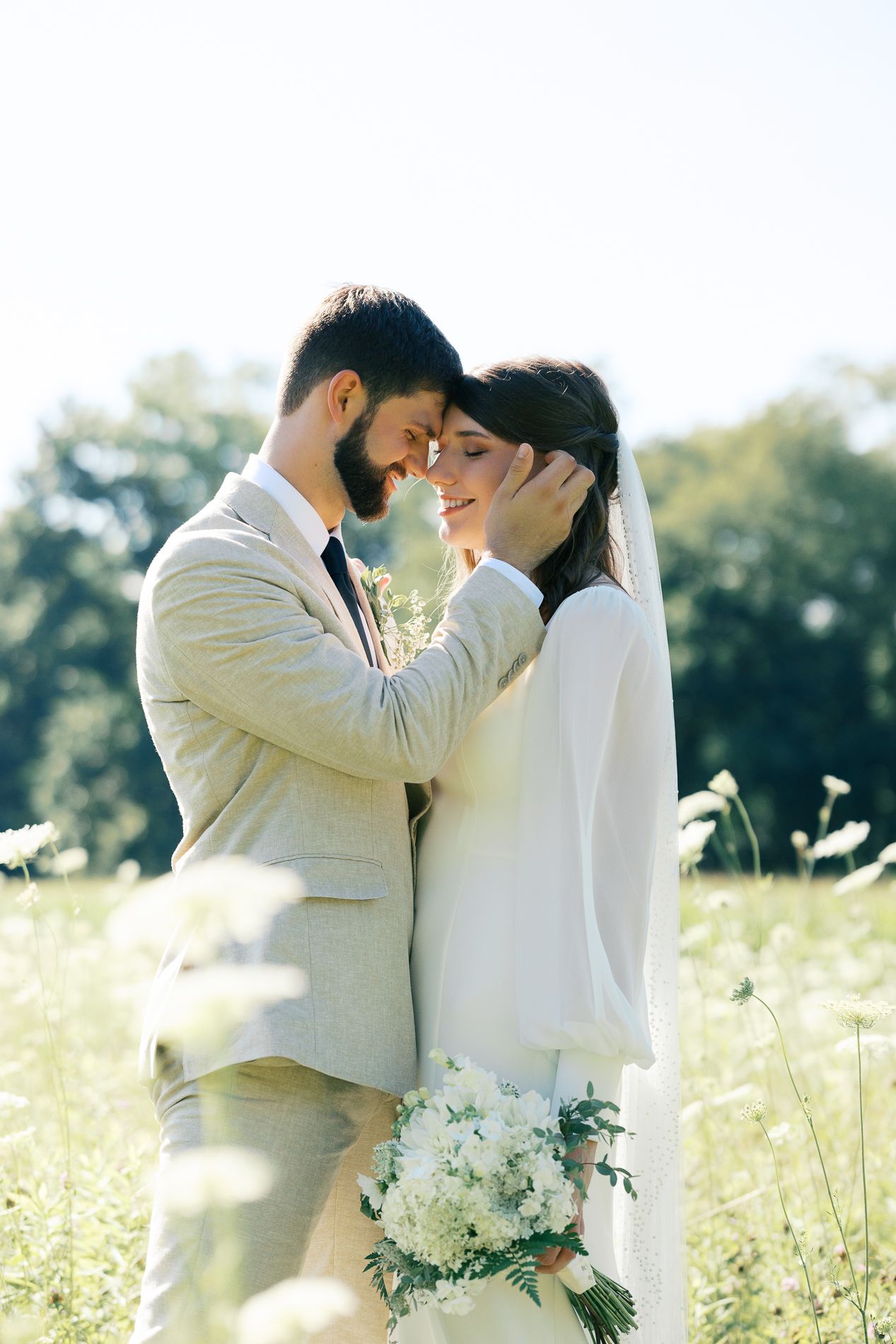 A bride and groom share a tender moment in a sunlit meadow, with the groom gently cradling her face.