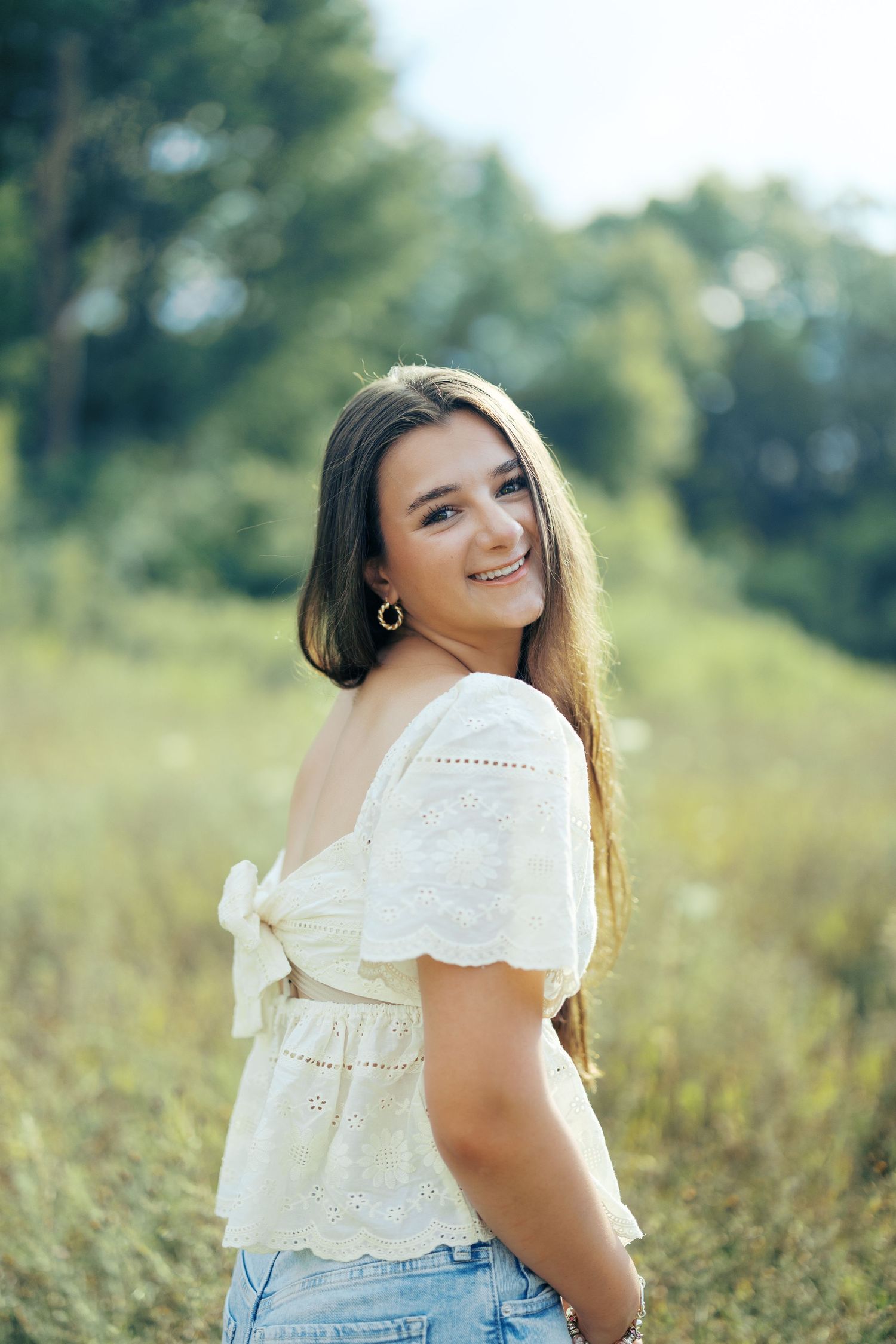 A person in a white eyelet lace top poses outdoors in a sunny field with natural lighting and bokeh effect.