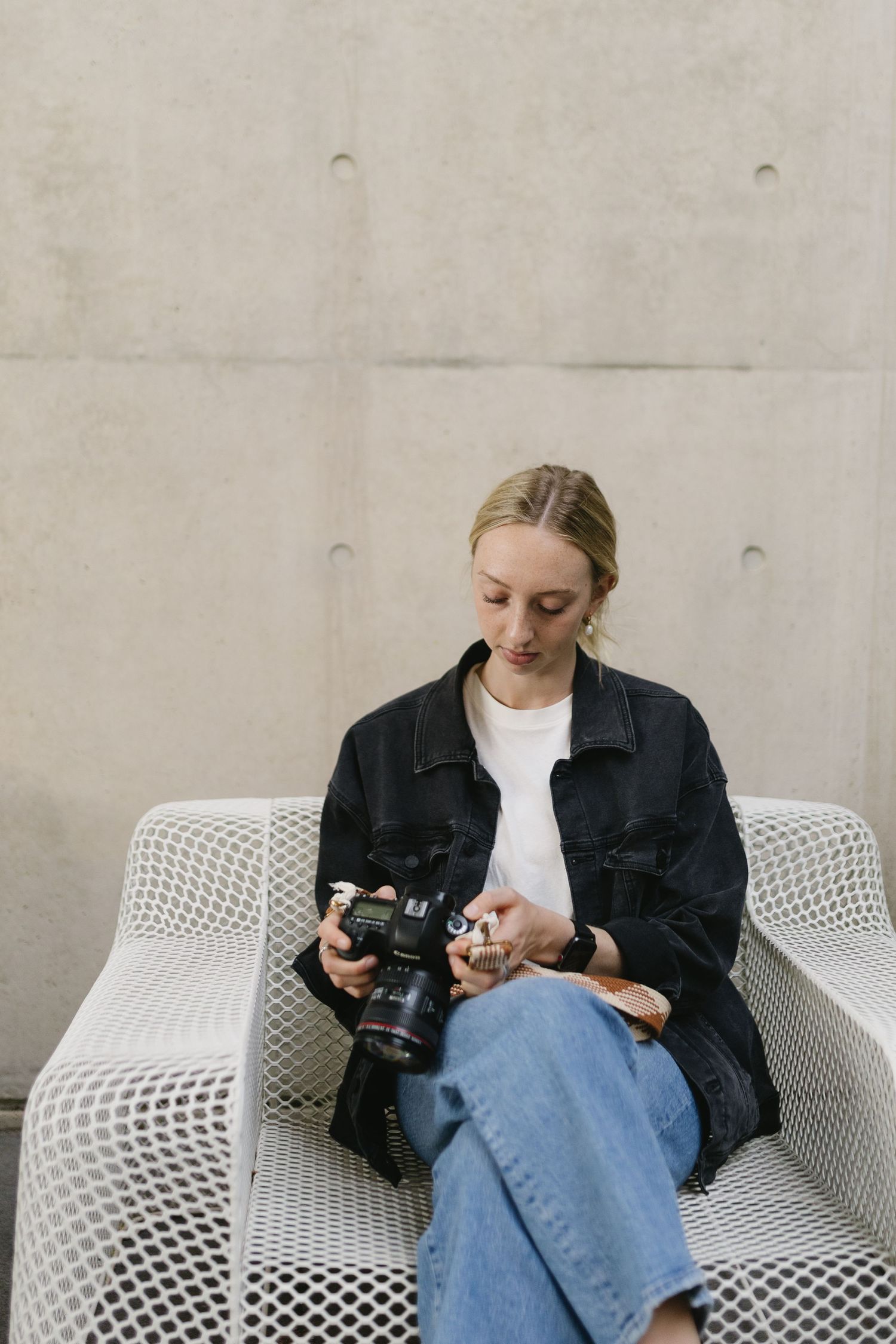 Photographer in denim jacket checking camera while sitting on modern white mesh chair.