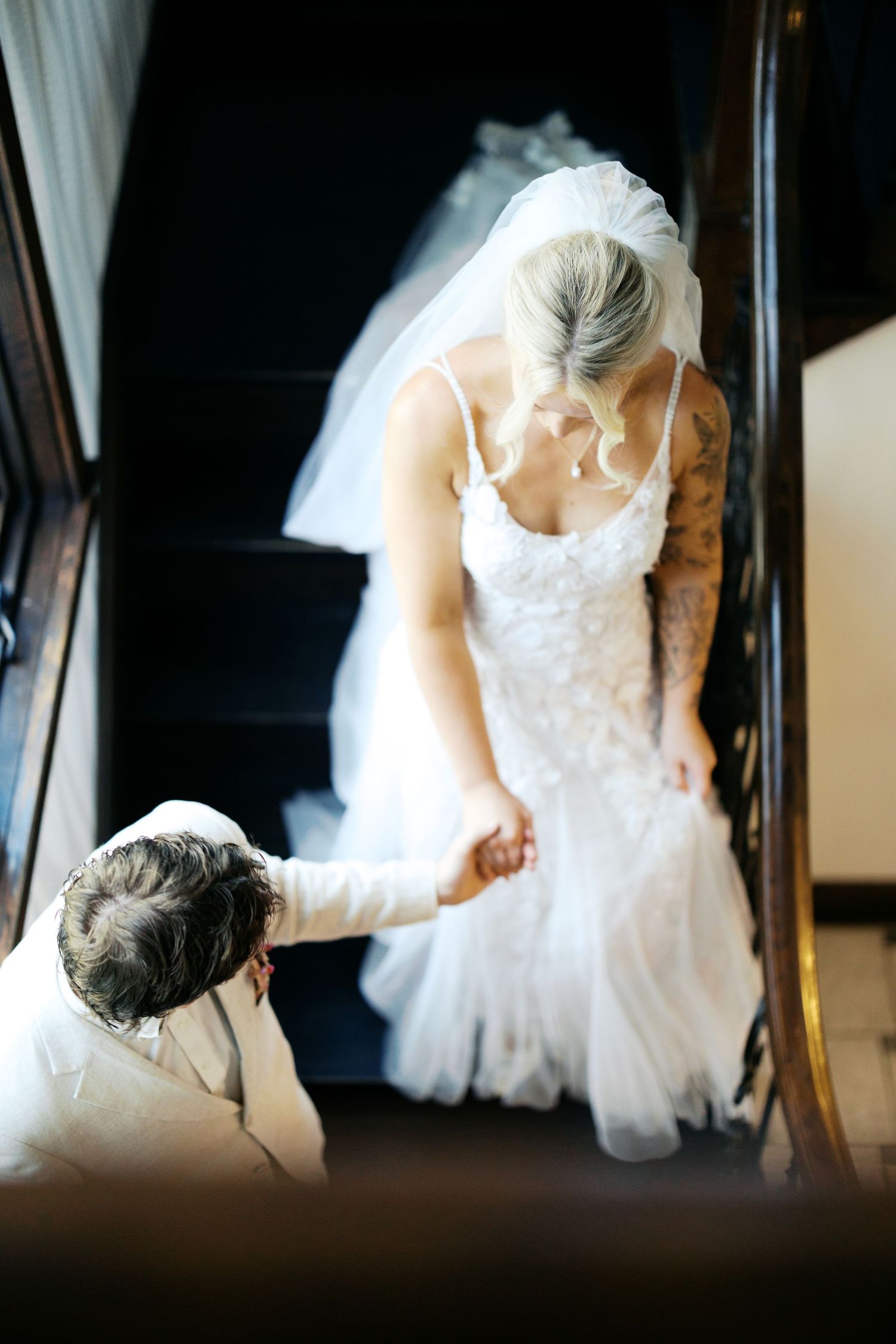 Groom kneeling on stairs helping bride with her white lace wedding dress and veil in elegant venue.