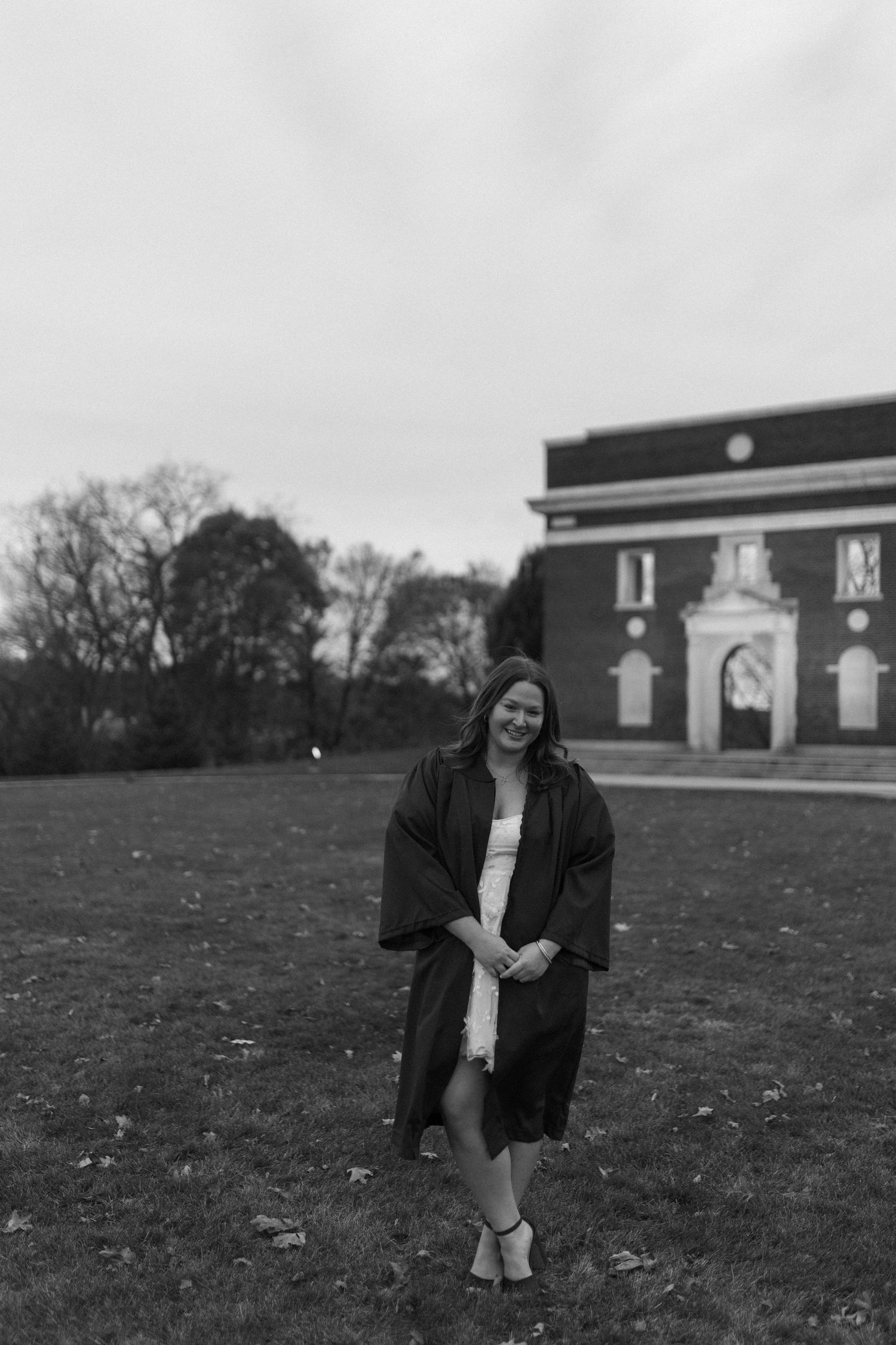 A black and white photo of an academic standing on grass in graduation attire with a brick building in the background.