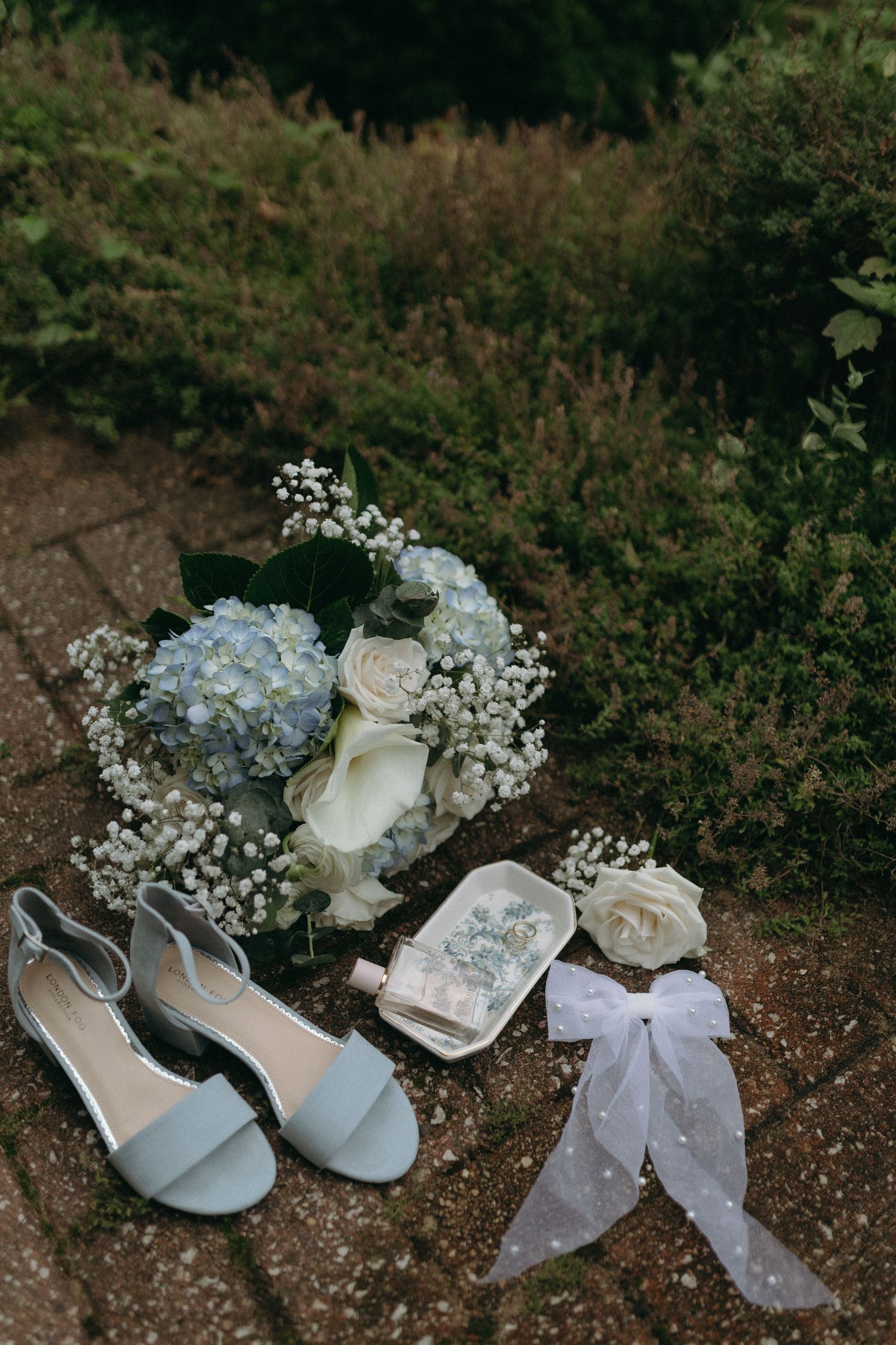 Wedding accessories including blue shoes, white flowers, and a lace bow arranged on ground.