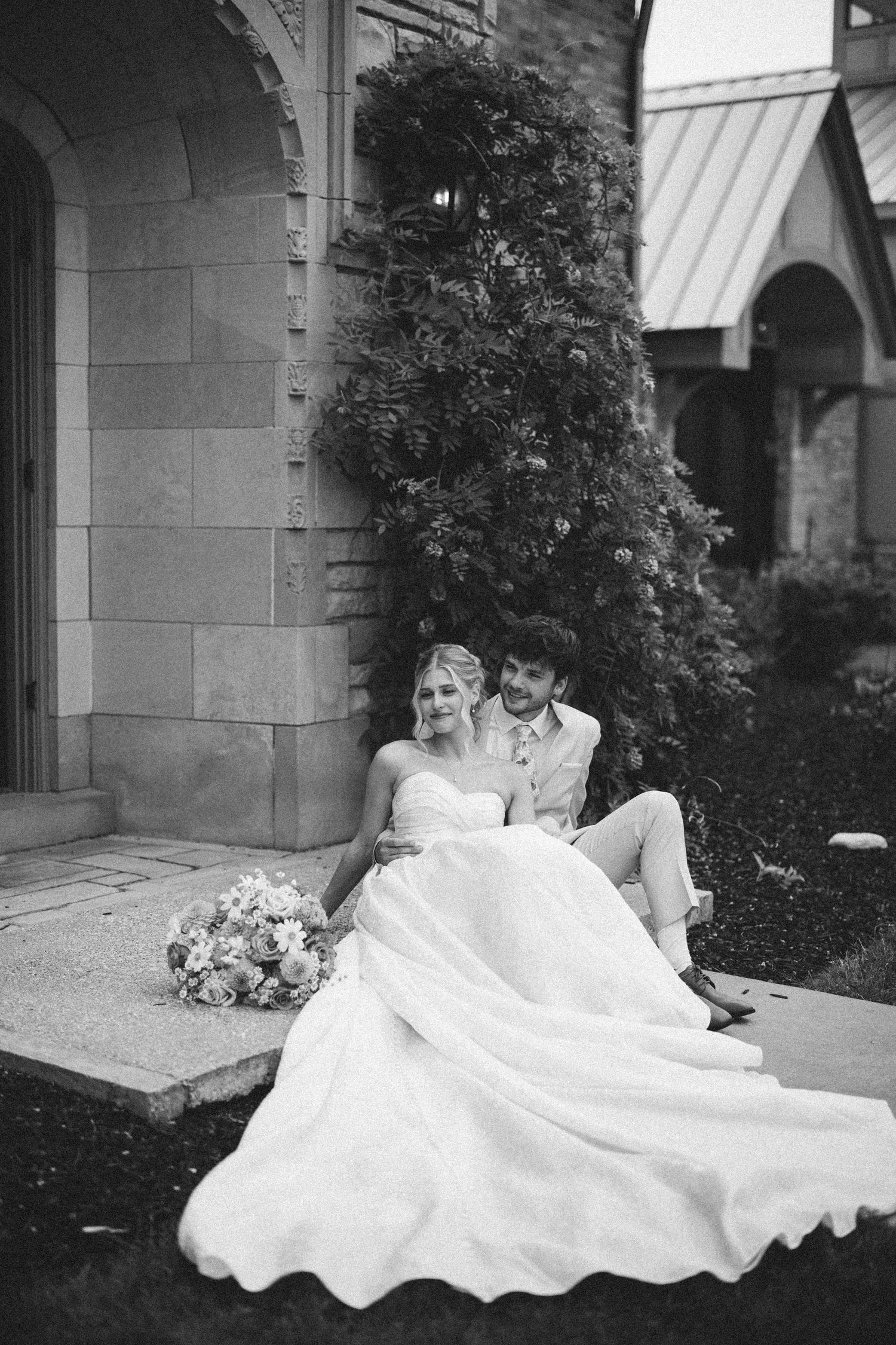 Black and white photo of a bride and groom sitting together outdoors near a stone building with a large evergreen tree.