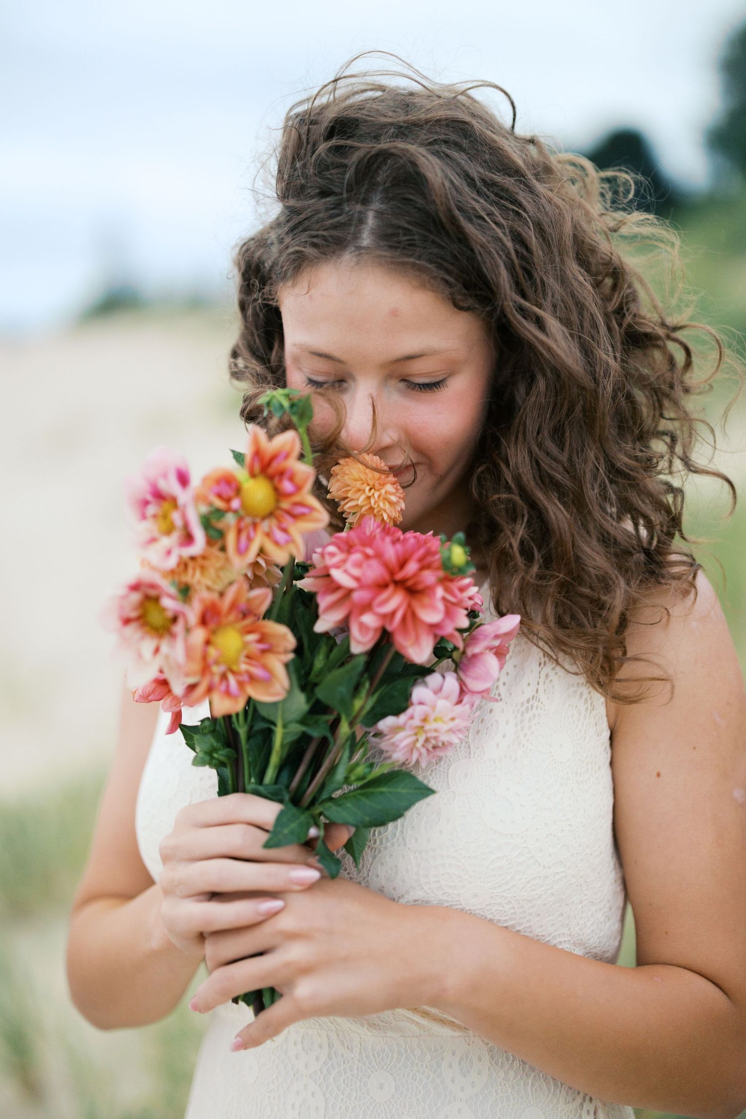 Young woman with curly hair smelling a bouquet of pink and orange dahlias outdoors.