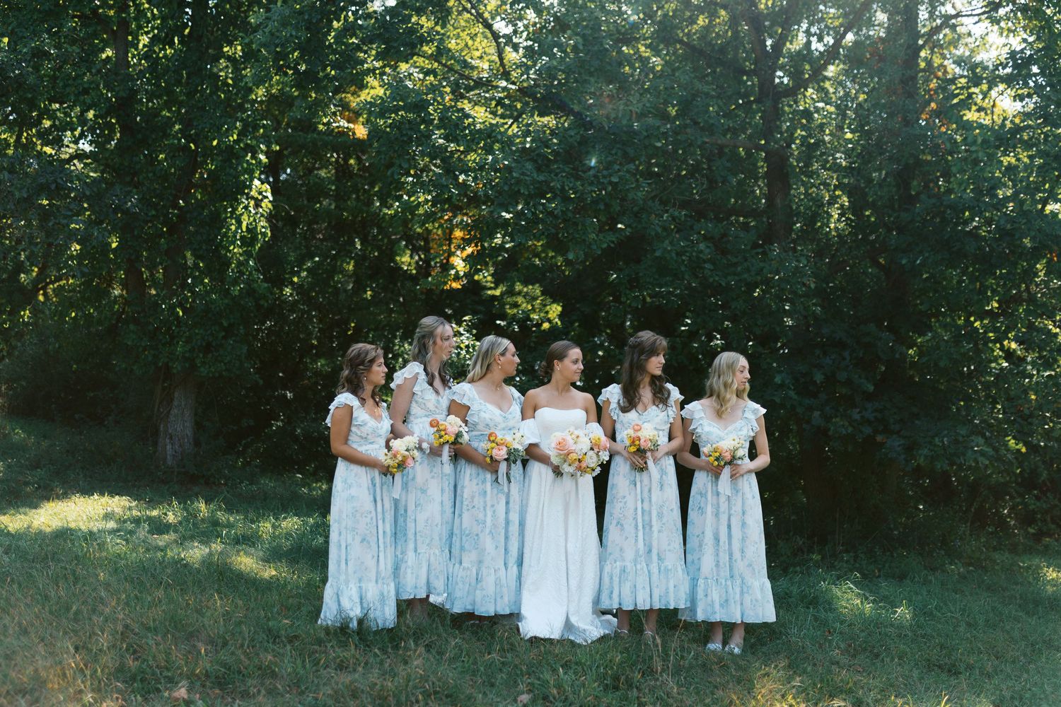 Bride and bridesmaids in floral blue dresses holding bouquets standing outdoors near lush green trees.