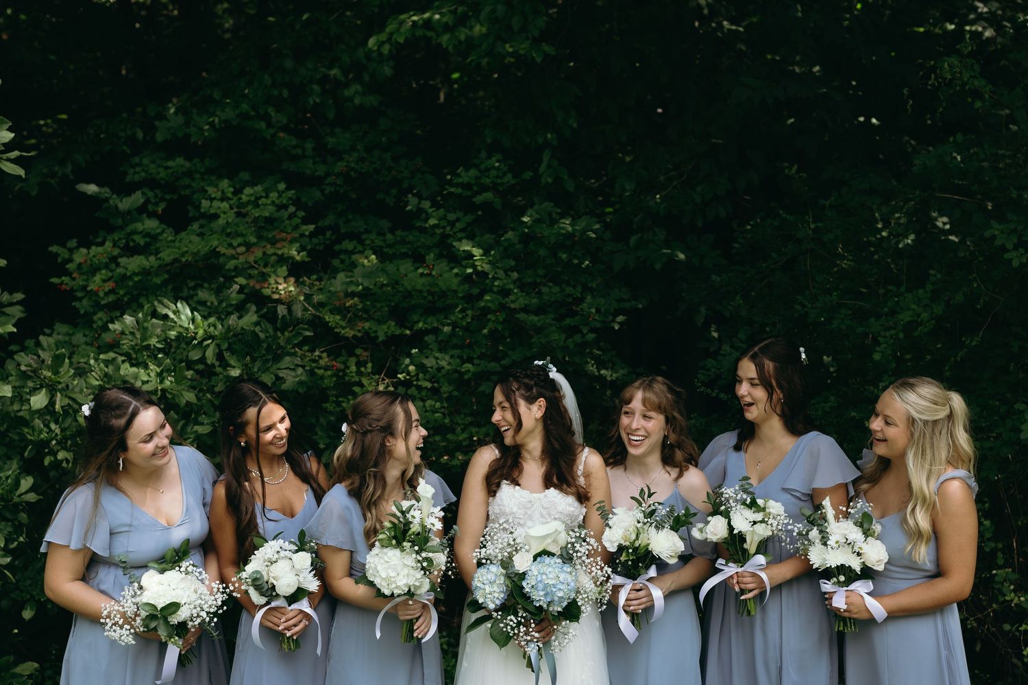 Bridesmaids in matching blue dresses laugh together while holding white floral bouquets.