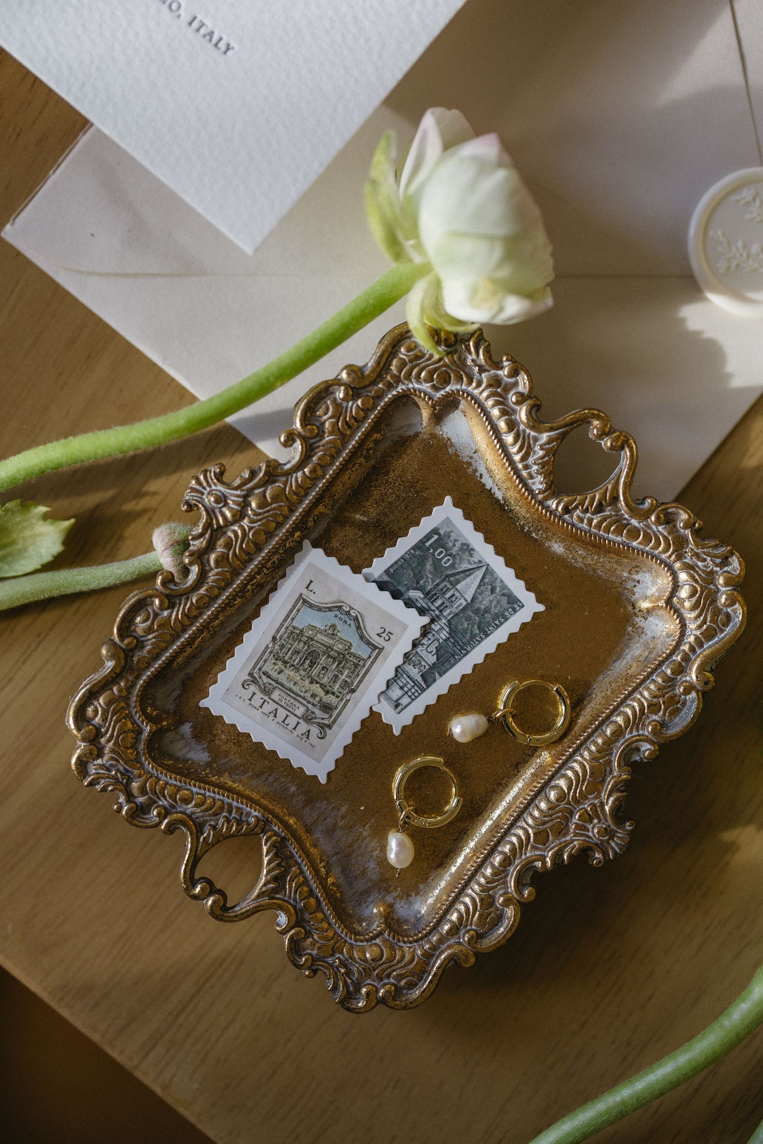Vintage ornate silver tray displays wedding rings and postage stamps alongside a white tulip on wooden surface.