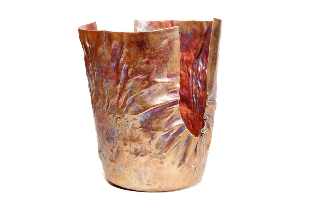 A hand formed copper vessel with wrinkle surface texture near the edge. With a U shape design cut down two opposing sides.